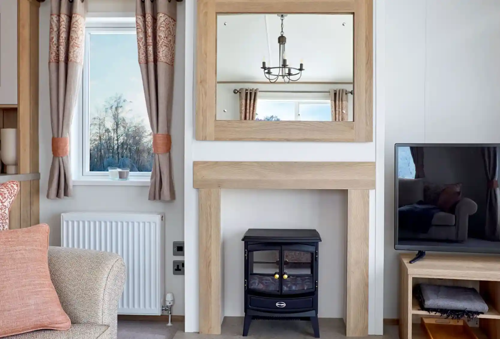 Stylish lounge area in the ABI Ambleside holiday home, featuring a wood-framed mirror above a modern fireplace with an electric log-effect stove, flanked by a window with patterned curtains and a flat-screen TV on a wooden media unit.