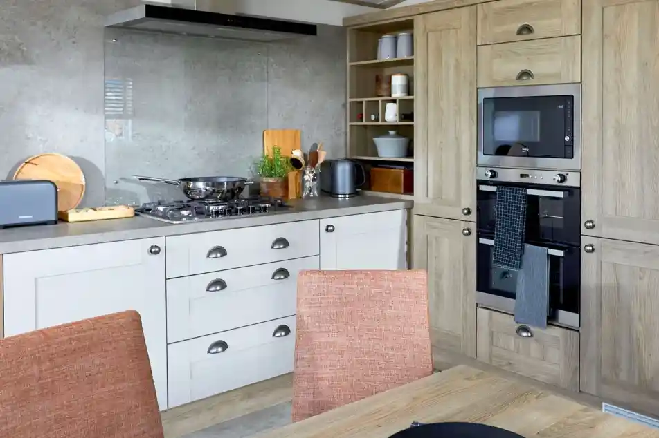 Modern kitchen in the ABI Ambleside holiday home featuring a built-in oven and microwave, integrated storage cabinets in a natural wood finish, gas hob with glass splashback and extractor fan, and a dining table with orange upholstered chairs in the foreground.