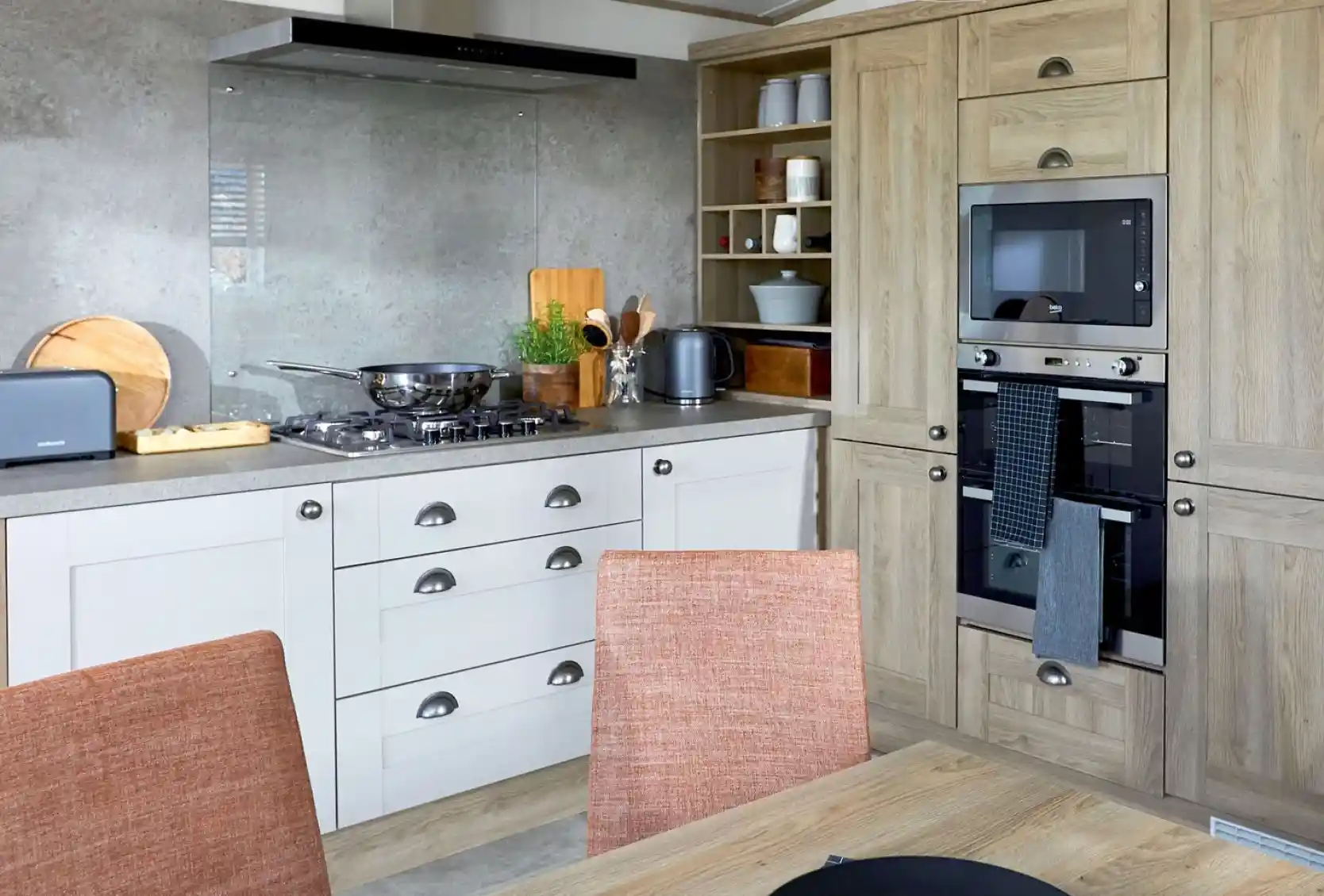 Modern kitchen in the ABI Ambleside holiday home featuring a built-in oven and microwave, integrated storage cabinets in a natural wood finish, gas hob with glass splashback and extractor fan, and a dining table with orange upholstered chairs in the foreground.