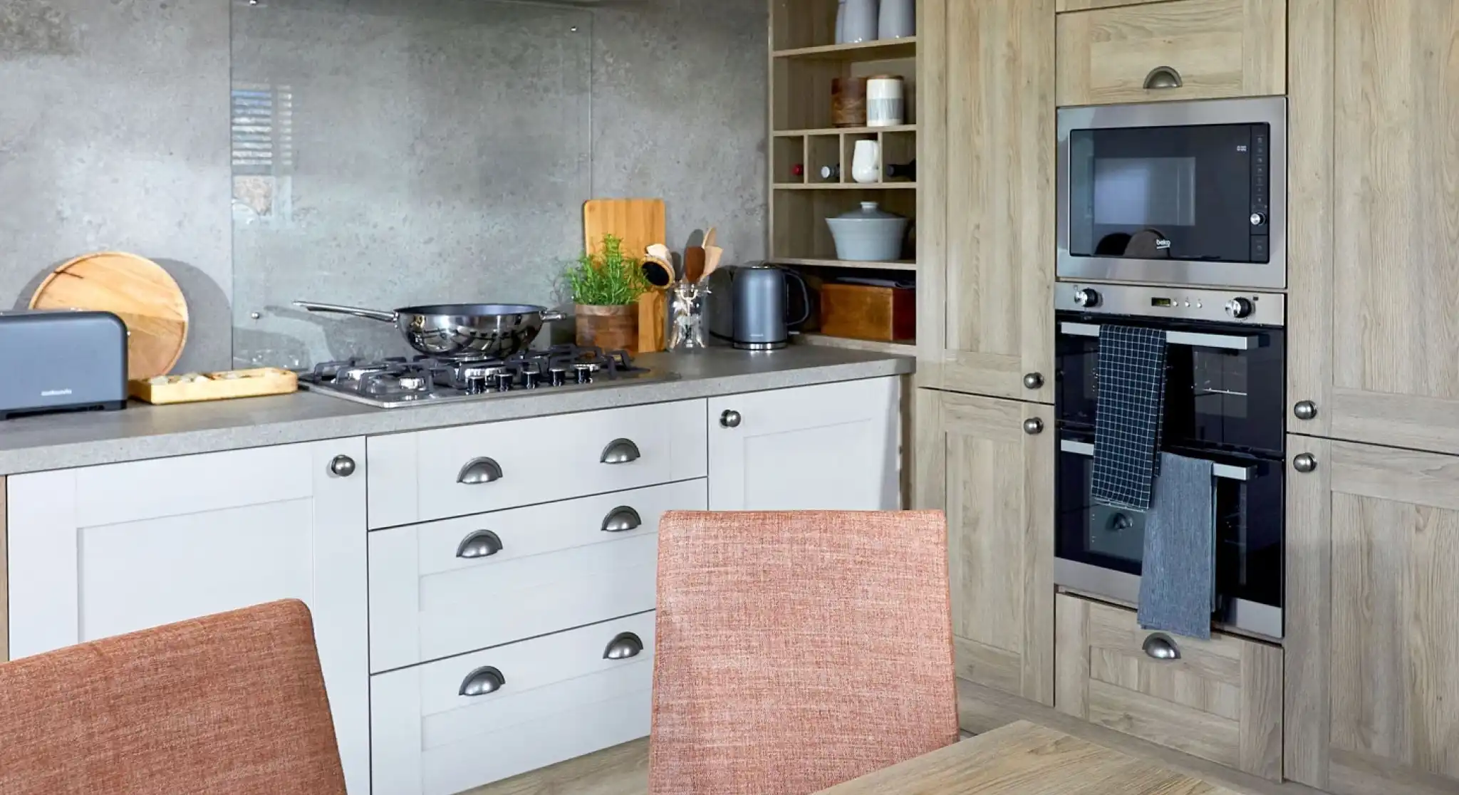 Modern kitchen in the ABI Ambleside holiday home featuring a built-in oven and microwave, integrated storage cabinets in a natural wood finish, gas hob with glass splashback and extractor fan, and a dining table with orange upholstered chairs in the foreground.