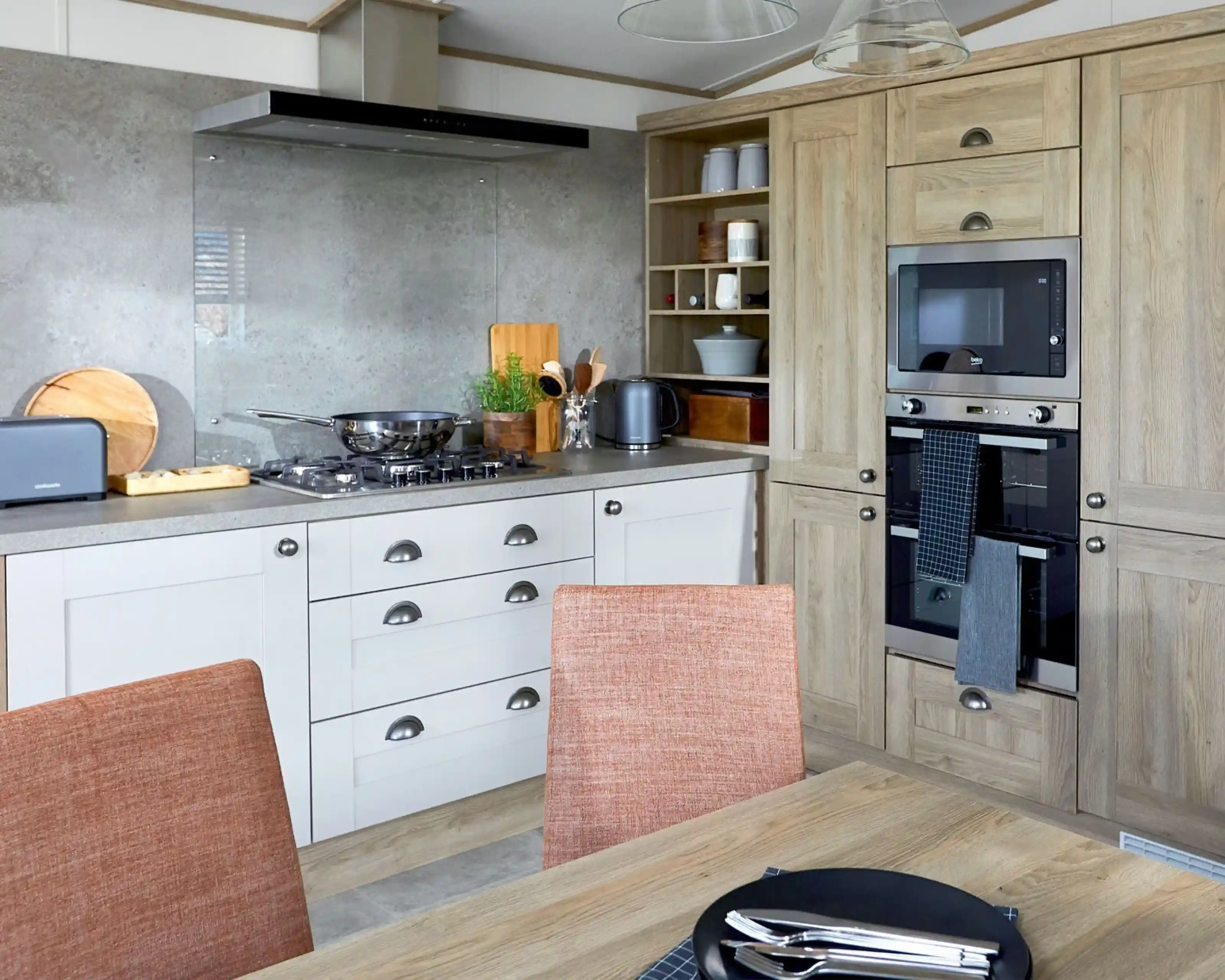 Modern kitchen in the ABI Ambleside holiday home featuring a built-in oven and microwave, integrated storage cabinets in a natural wood finish, gas hob with glass splashback and extractor fan, and a dining table with orange upholstered chairs in the foreground.