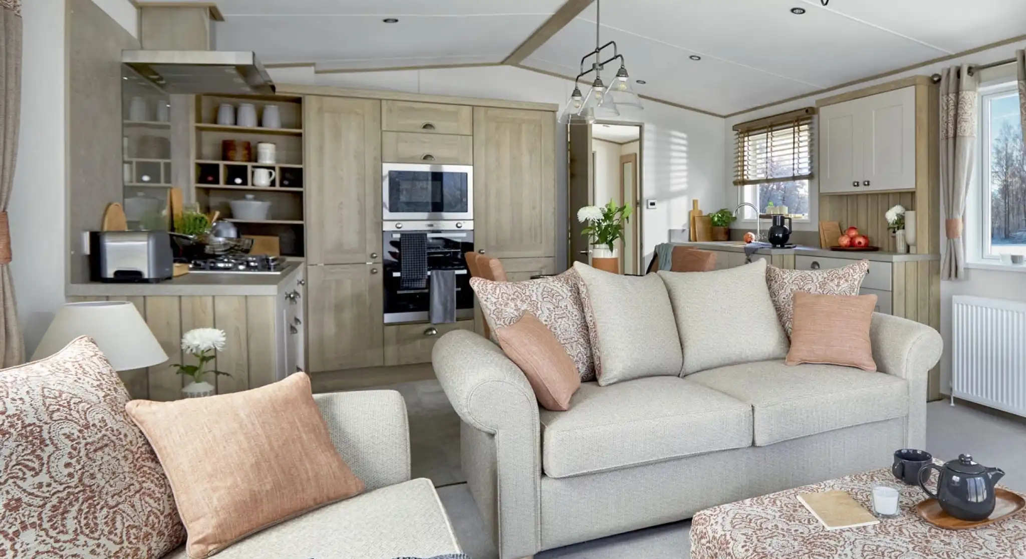 Open-plan lounge and kitchen area of the ABI Ambleside holiday home, featuring a neutral-toned sofa with patterned cushions, a matching upholstered footstool with a tea tray, a modern fitted kitchen with integrated appliances, and large windows letting in natural light.