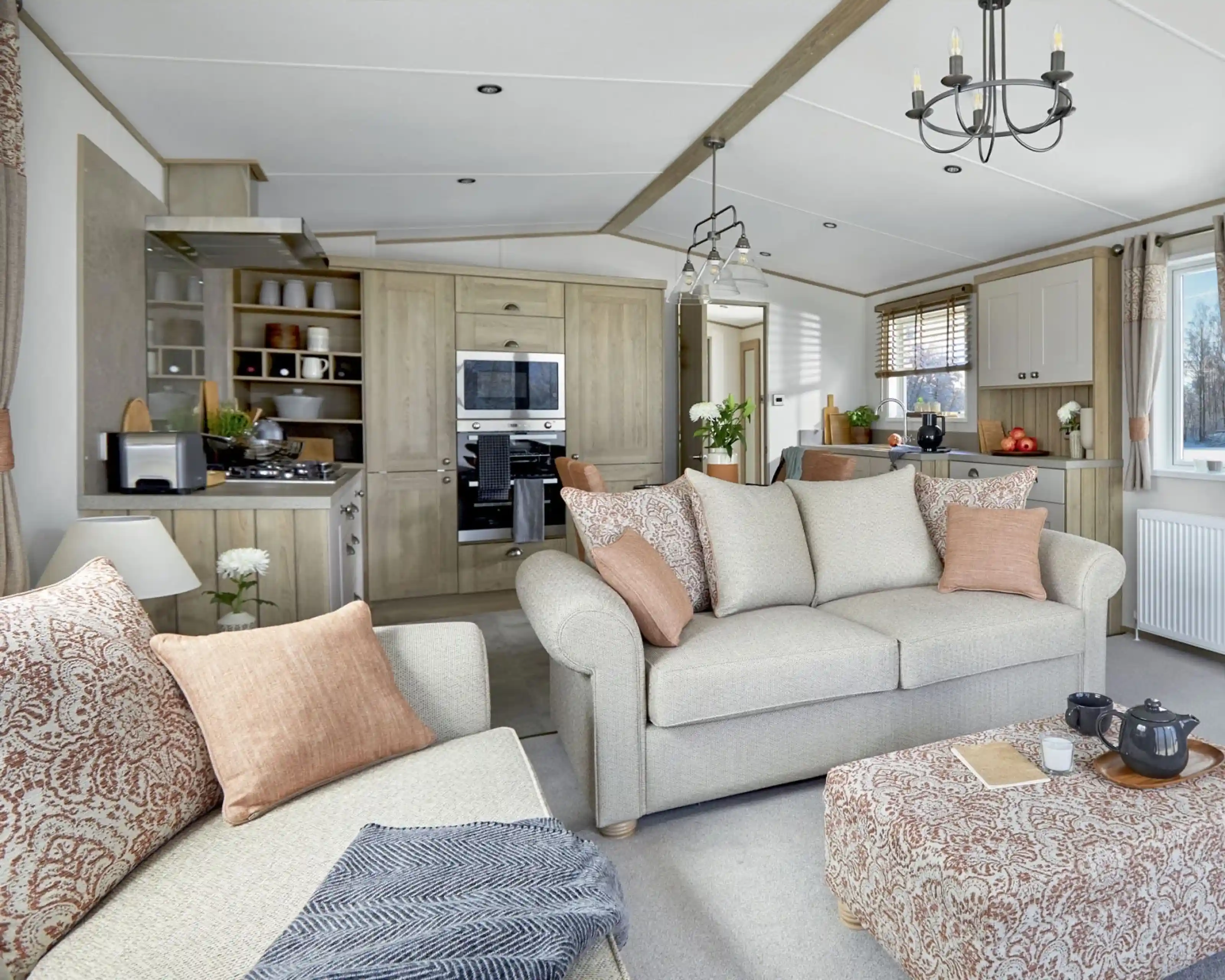 Open-plan lounge and kitchen area of the ABI Ambleside holiday home, featuring a neutral-toned sofa with patterned cushions, a matching upholstered footstool with a tea tray, a modern fitted kitchen with integrated appliances, and large windows letting in natural light.