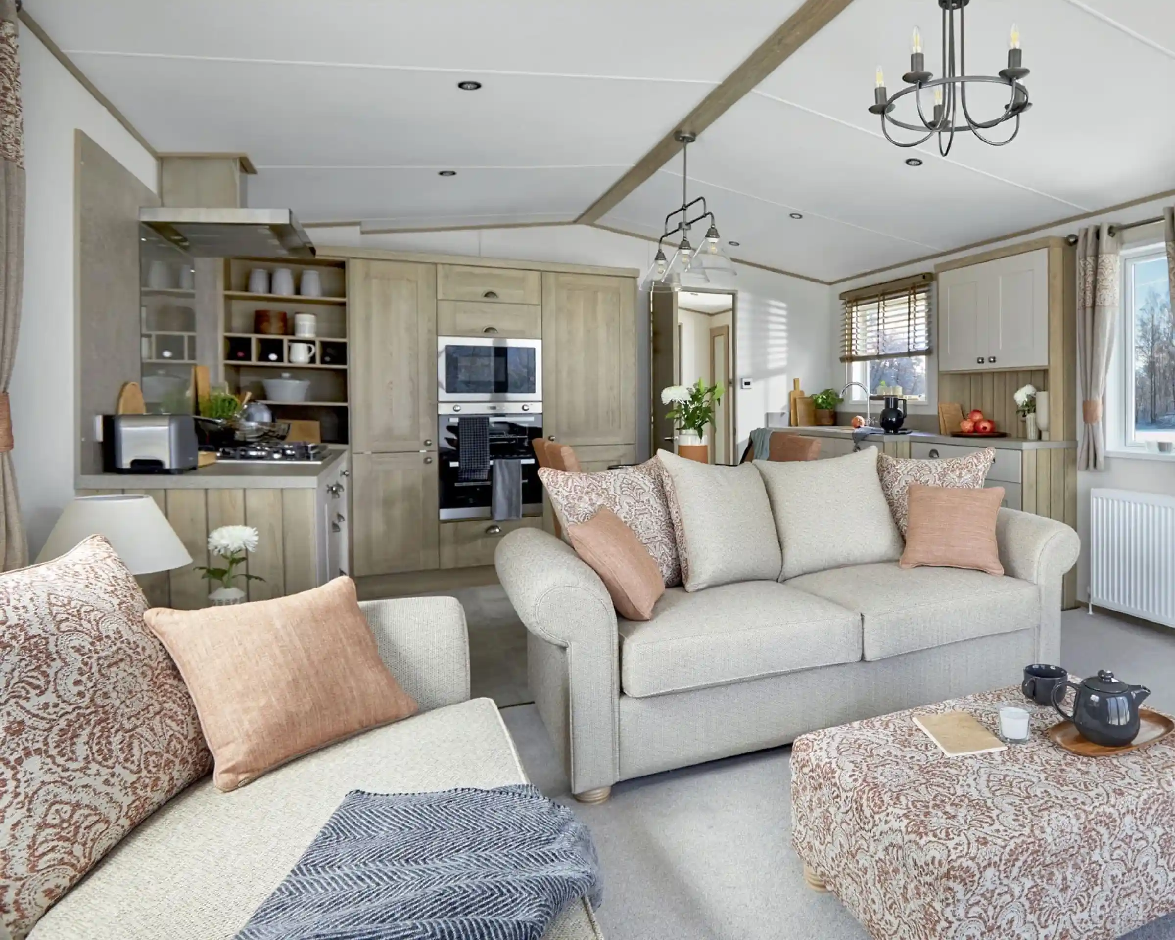 Open-plan lounge and kitchen area of the ABI Ambleside holiday home, featuring a neutral-toned sofa with patterned cushions, a matching upholstered footstool with a tea tray, a modern fitted kitchen with integrated appliances, and large windows letting in natural light.