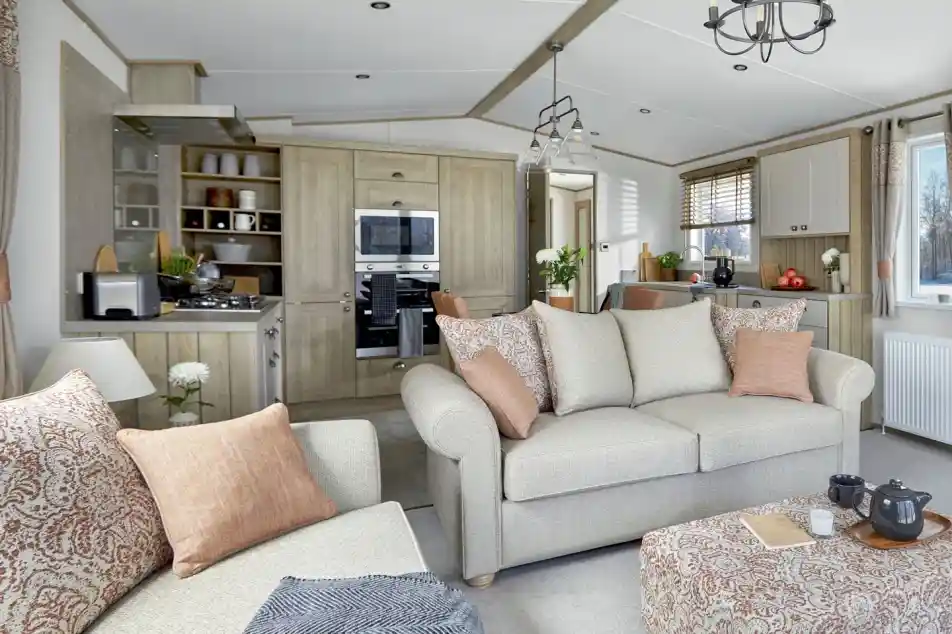 Open-plan lounge and kitchen area of the ABI Ambleside holiday home, featuring a neutral-toned sofa with patterned cushions, a matching upholstered footstool with a tea tray, a modern fitted kitchen with integrated appliances, and large windows letting in natural light.