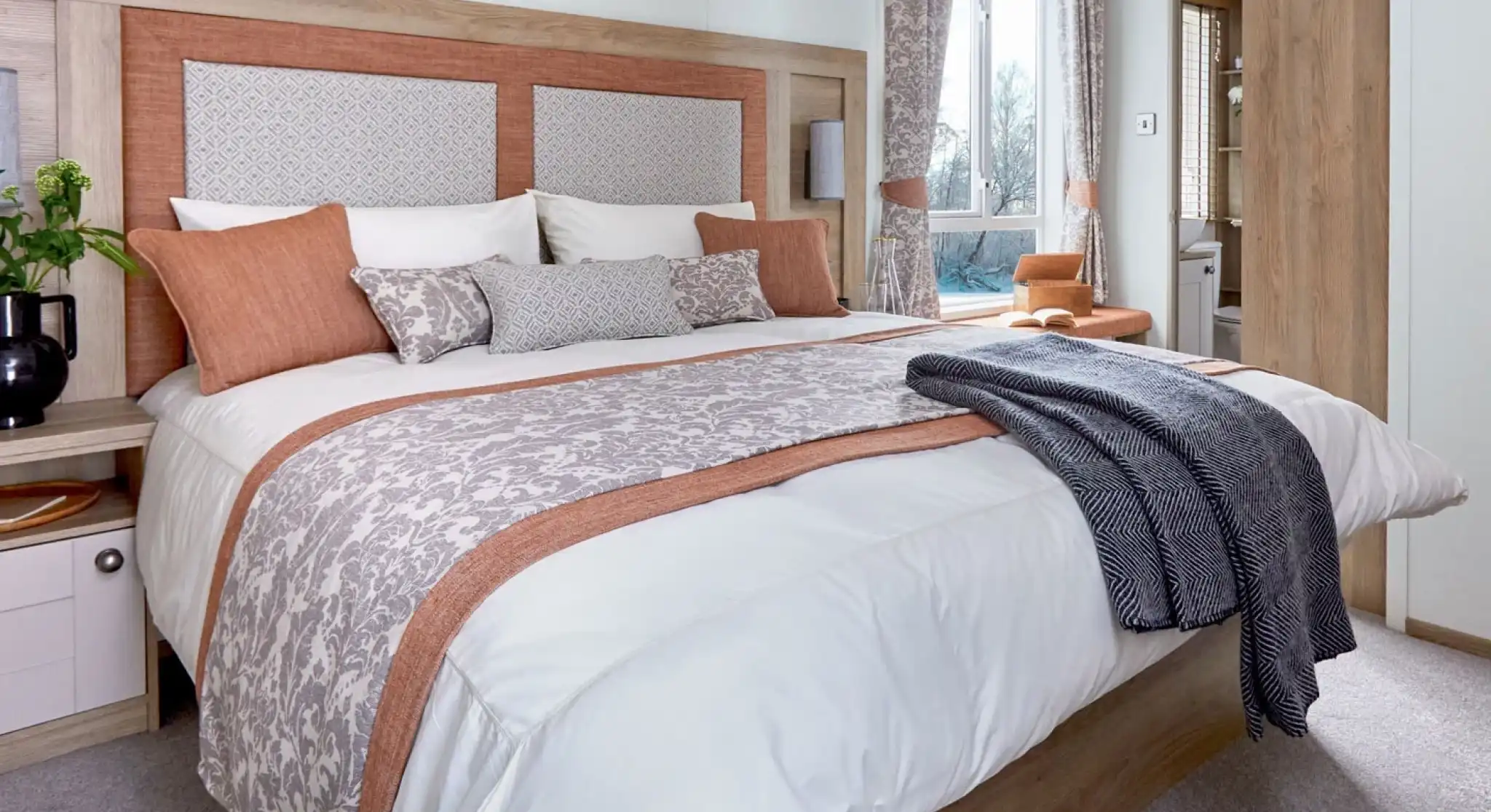 A neatly made bed with white and patterned beige bedding accented by burnt orange pillows and trim. The wooden headboard has padded panels, and a dark gray throw blanket is draped over the foot of the bed. A window with matching curtains and a small bedside table with a plant are visible in the bright, cozy bedroom.
