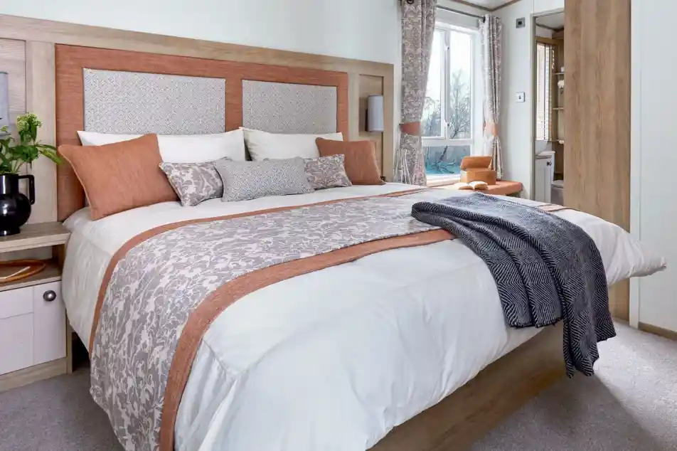 A neatly made bed with white and patterned beige bedding accented by burnt orange pillows and trim. The wooden headboard has padded panels, and a dark gray throw blanket is draped over the foot of the bed. A window with matching curtains and a small bedside table with a plant are visible in the bright, cozy bedroom.