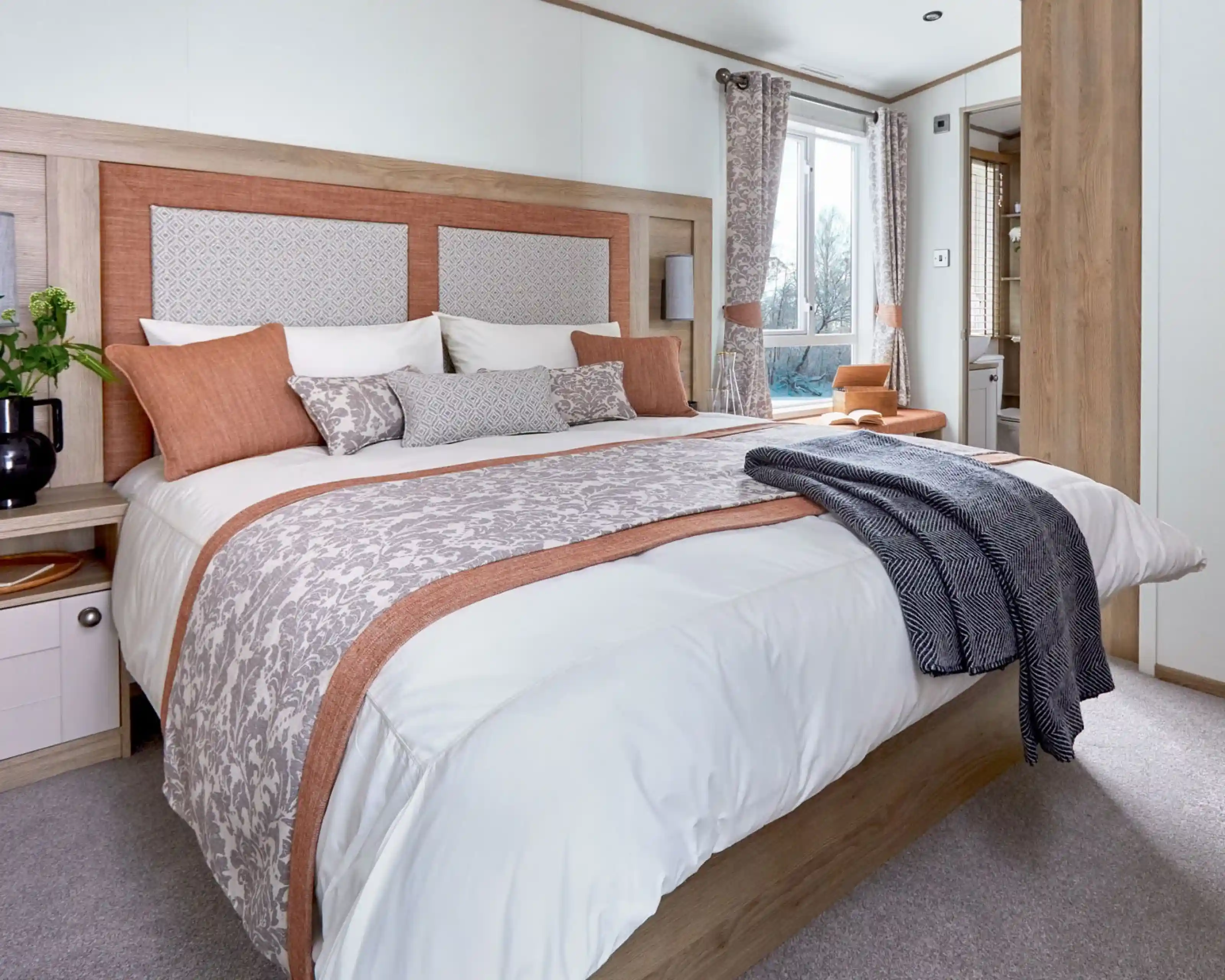 A neatly made bed with white and patterned beige bedding accented by burnt orange pillows and trim. The wooden headboard has padded panels, and a dark gray throw blanket is draped over the foot of the bed. A window with matching curtains and a small bedside table with a plant are visible in the bright, cozy bedroom.