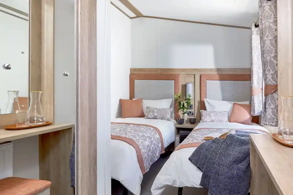 A cozy twin bedroom with two single beds, each dressed in white linens with decorative pillows and patterned throws in soft peach and gray tones. A small wooden nightstand with two mugs and a vase with green foliage sits between the beds. Light wood furniture, including a dressing table with a mirror and a matching stool with a peach cushion, complements the room. Patterned curtains in gray and peach hang by the window, and a dark textured blanket is draped over one of the beds. The room has a warm, inviting atmosphere with neutral and pastel colors.