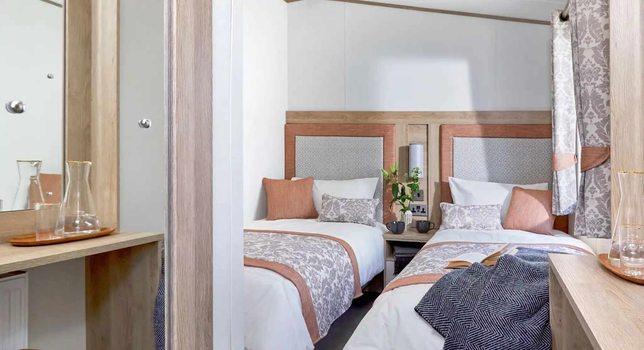 A cozy twin bedroom with two single beds, each dressed in white linens with decorative pillows and patterned throws in soft peach and gray tones. A small wooden nightstand with two mugs and a vase with green foliage sits between the beds. Light wood furniture, including a dressing table with a mirror and a matching stool with a peach cushion, complements the room. Patterned curtains in gray and peach hang by the window, and a dark textured blanket is draped over one of the beds. The room has a warm, inviting atmosphere with neutral and pastel colors.