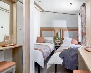 A cozy twin bedroom with two single beds, each dressed in white linens with decorative pillows and patterned throws in soft peach and gray tones. A small wooden nightstand with two mugs and a vase with green foliage sits between the beds. Light wood furniture, including a dressing table with a mirror and a matching stool with a peach cushion, complements the room. Patterned curtains in gray and peach hang by the window, and a dark textured blanket is draped over one of the beds. The room has a warm, inviting atmosphere with neutral and pastel colors.