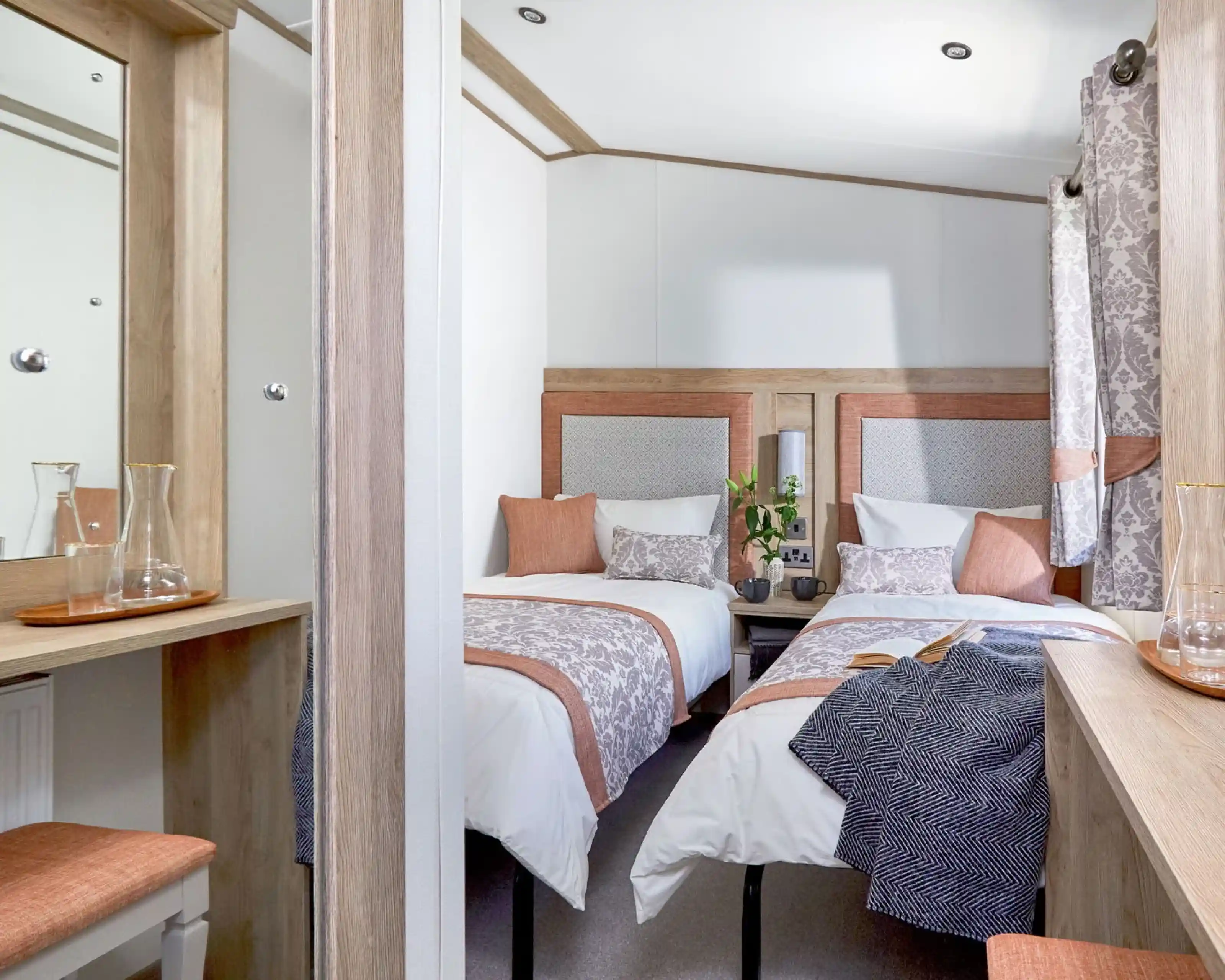 A cozy twin bedroom with two single beds, each dressed in white linens with decorative pillows and patterned throws in soft peach and gray tones. A small wooden nightstand with two mugs and a vase with green foliage sits between the beds. Light wood furniture, including a dressing table with a mirror and a matching stool with a peach cushion, complements the room. Patterned curtains in gray and peach hang by the window, and a dark textured blanket is draped over one of the beds. The room has a warm, inviting atmosphere with neutral and pastel colors.