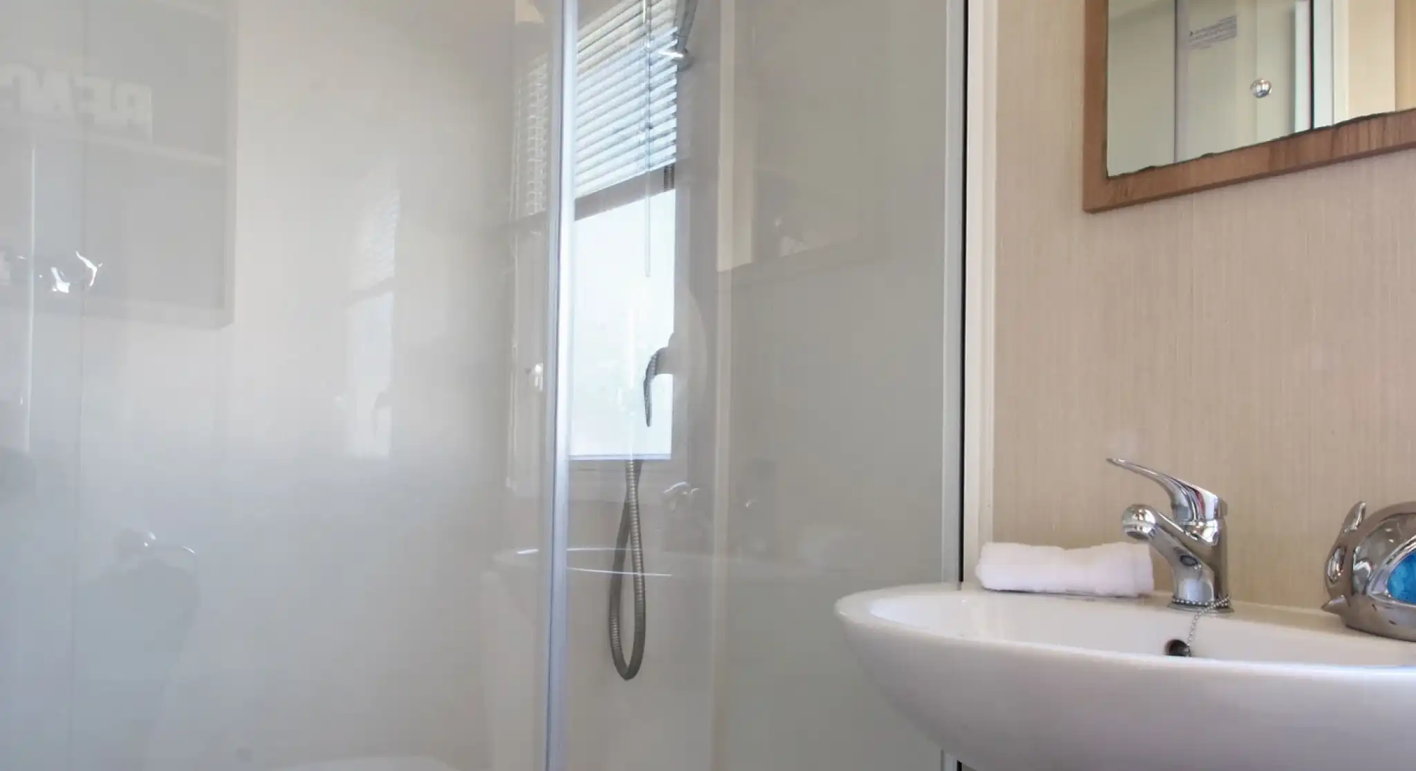 This image shows a clean and functional bathroom, featuring a compact shower area with a glass partition and a small washbasin. The shower has a modern overhead showerhead and a handheld shower attachment, offering flexibility. The sink area includes a faucet with a towel neatly placed next to it. A mirror is mounted above the sink, adding to the sense of openness in the space. The bathroom is well-lit, with natural light streaming in through a window with blinds. The overall design is minimalistic, contributing to a fresh and organized atmosphere.