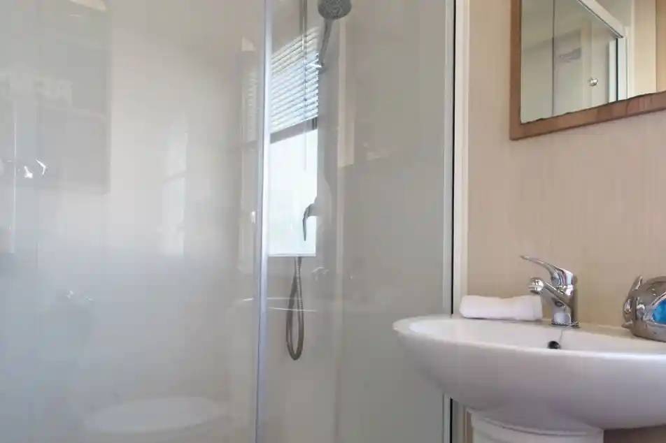 This image shows a clean and functional bathroom, featuring a compact shower area with a glass partition and a small washbasin. The shower has a modern overhead showerhead and a handheld shower attachment, offering flexibility. The sink area includes a faucet with a towel neatly placed next to it. A mirror is mounted above the sink, adding to the sense of openness in the space. The bathroom is well-lit, with natural light streaming in through a window with blinds. The overall design is minimalistic, contributing to a fresh and organized atmosphere.