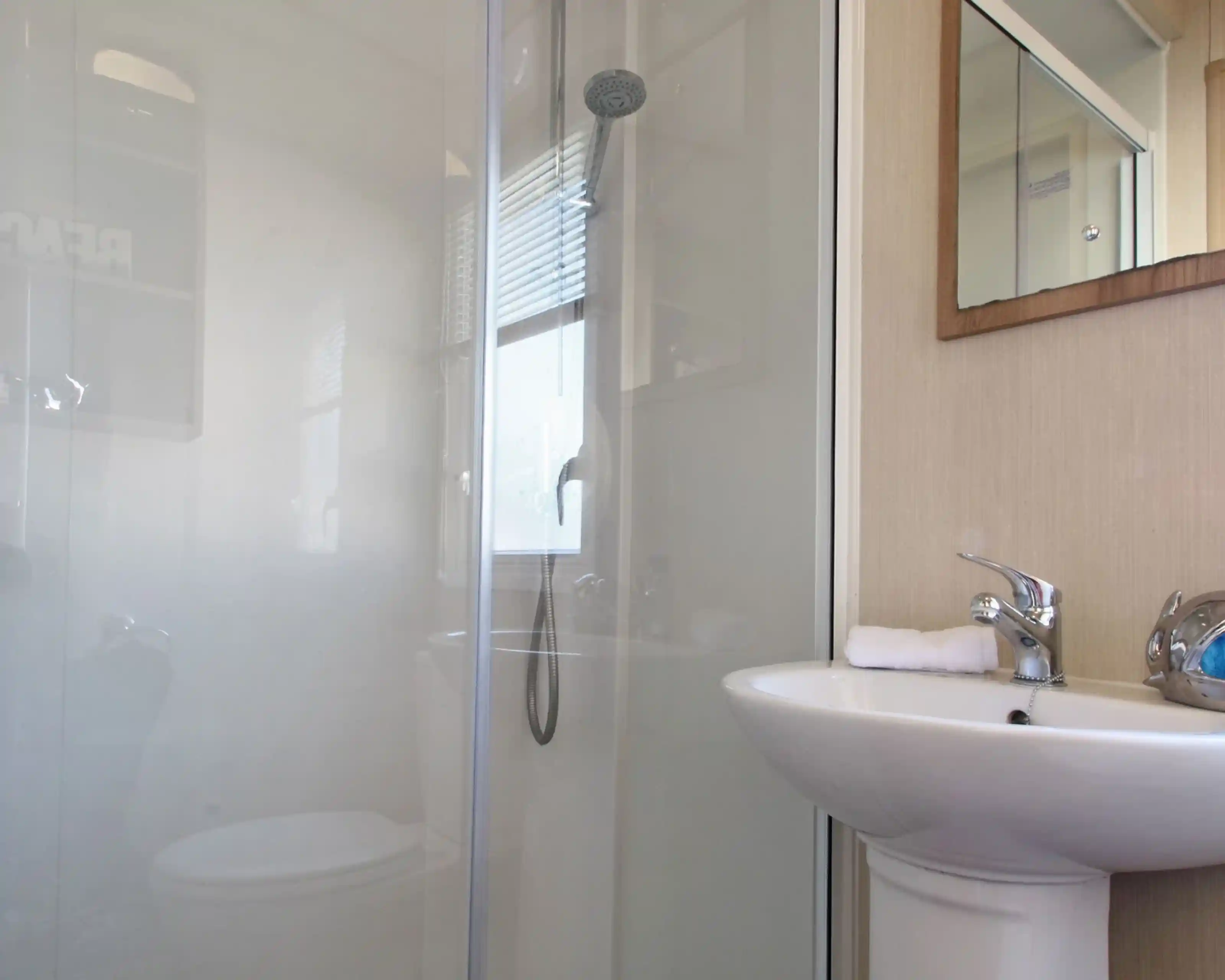 This image shows a clean and functional bathroom, featuring a compact shower area with a glass partition and a small washbasin. The shower has a modern overhead showerhead and a handheld shower attachment, offering flexibility. The sink area includes a faucet with a towel neatly placed next to it. A mirror is mounted above the sink, adding to the sense of openness in the space. The bathroom is well-lit, with natural light streaming in through a window with blinds. The overall design is minimalistic, contributing to a fresh and organized atmosphere.
