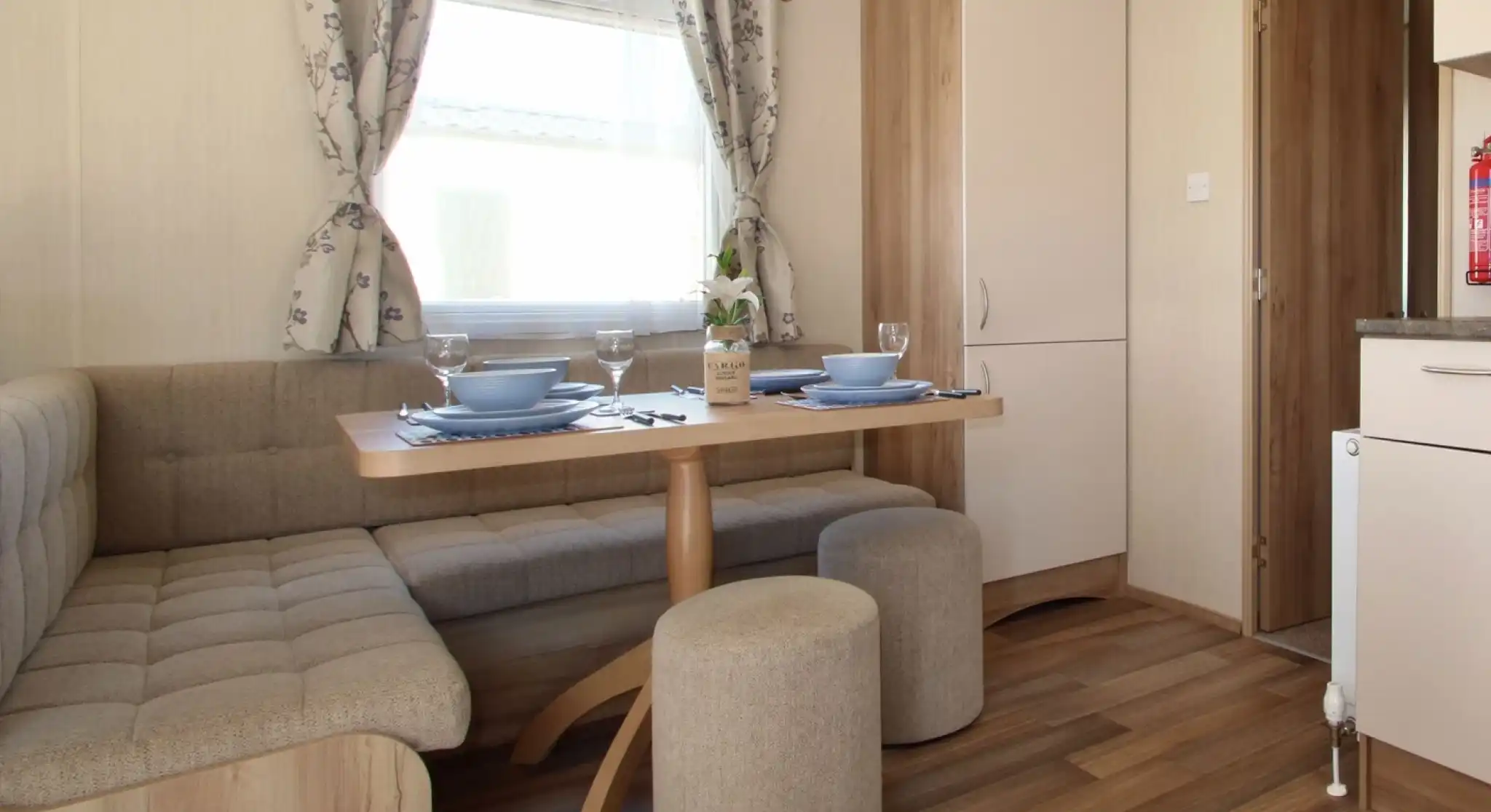 This image showcases a cozy and functional dining nook. The space features a built-in bench with soft cushions around a light wood table, creating a comfortable area for dining or relaxing. There are two matching stools for extra seating, and the table is set with blue plates, glasses, and a simple vase with a flower. The room has large windows dressed in soft floral curtains that let in natural light, and the overall design feels welcoming and bright. The adjacent kitchen area is visible through the doorway, adding to the open, integrated feel of the space.