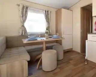 This image showcases a cozy and functional dining nook. The space features a built-in bench with soft cushions around a light wood table, creating a comfortable area for dining or relaxing. There are two matching stools for extra seating, and the table is set with blue plates, glasses, and a simple vase with a flower. The room has large windows dressed in soft floral curtains that let in natural light, and the overall design feels welcoming and bright. The adjacent kitchen area is visible through the doorway, adding to the open, integrated feel of the space.