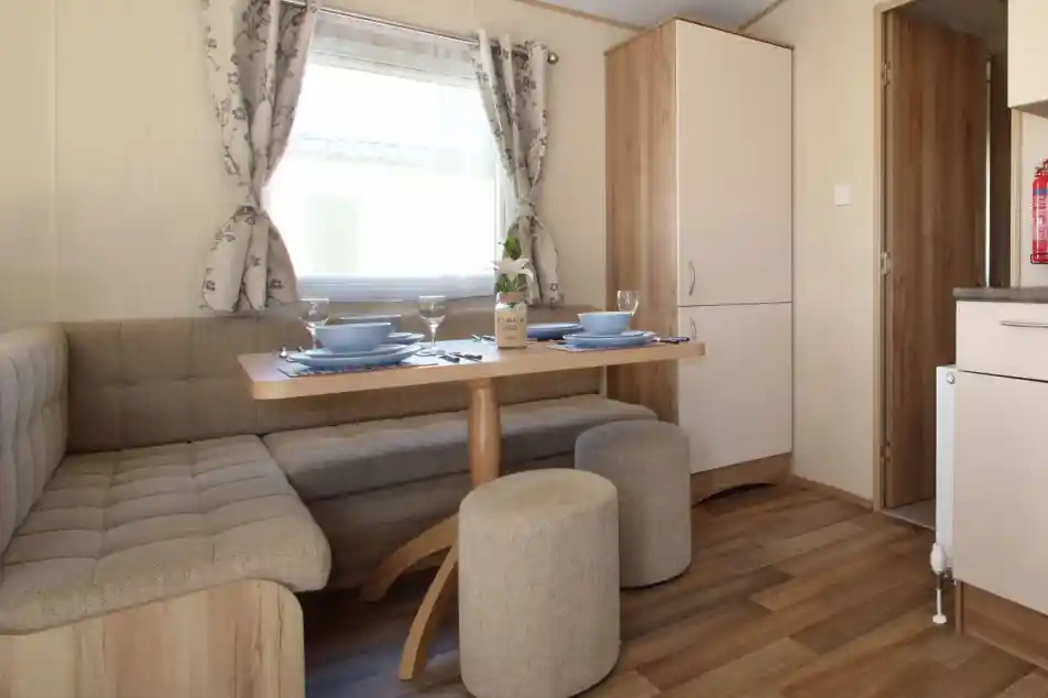 This image showcases a cozy and functional dining nook. The space features a built-in bench with soft cushions around a light wood table, creating a comfortable area for dining or relaxing. There are two matching stools for extra seating, and the table is set with blue plates, glasses, and a simple vase with a flower. The room has large windows dressed in soft floral curtains that let in natural light, and the overall design feels welcoming and bright. The adjacent kitchen area is visible through the doorway, adding to the open, integrated feel of the space.