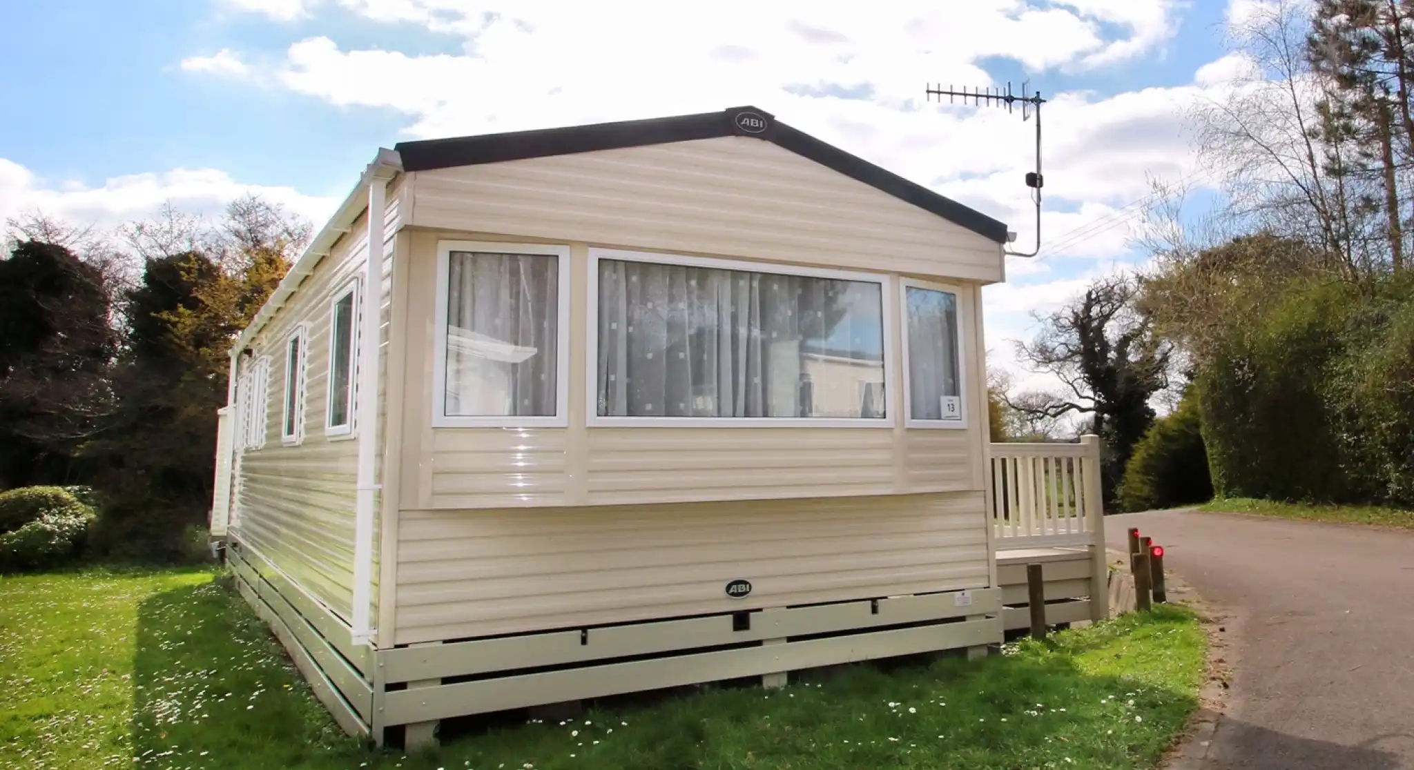 This image shows the exterior of a caravan or mobile home, set in a peaceful, green environment. The structure has light-colored siding with large windows, allowing plenty of natural light to enter. The small wooden deck at the entrance adds a welcoming touch. The surrounding grass is lush, with some wildflowers dotting the ground. The sky above is partly cloudy, and the trees along the road provide a scenic backdrop. The location appears to be tranquil and ideal for a relaxing stay.