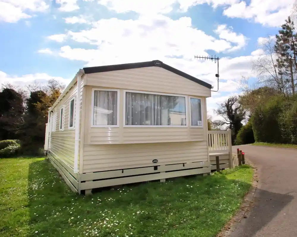 This image shows the exterior of a caravan or mobile home, set in a peaceful, green environment. The structure has light-colored siding with large windows, allowing plenty of natural light to enter. The small wooden deck at the entrance adds a welcoming touch. The surrounding grass is lush, with some wildflowers dotting the ground. The sky above is partly cloudy, and the trees along the road provide a scenic backdrop. The location appears to be tranquil and ideal for a relaxing stay.