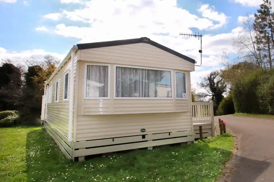 This image shows the exterior of a caravan or mobile home, set in a peaceful, green environment. The structure has light-colored siding with large windows, allowing plenty of natural light to enter. The small wooden deck at the entrance adds a welcoming touch. The surrounding grass is lush, with some wildflowers dotting the ground. The sky above is partly cloudy, and the trees along the road provide a scenic backdrop. The location appears to be tranquil and ideal for a relaxing stay.