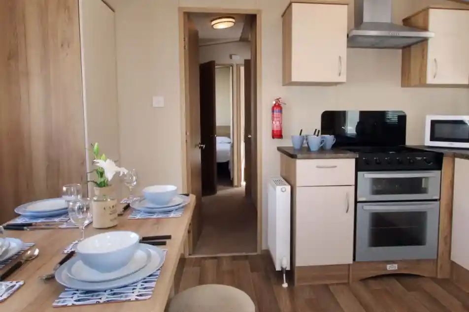 This image shows a clean, well-organized kitchen and dining area. The space is bright and functional, with light-colored cabinets and modern appliances, including an oven, microwave, and a fire extinguisher mounted on the wall. The dining table is set for a meal, with blue and white tableware and a simple vase with a flower adding a decorative touch. The flooring is wood-like laminate, contributing to a cozy and practical atmosphere. Through the open door, there’s a glimpse of a bedroom, showing a soft and comfortable environment.