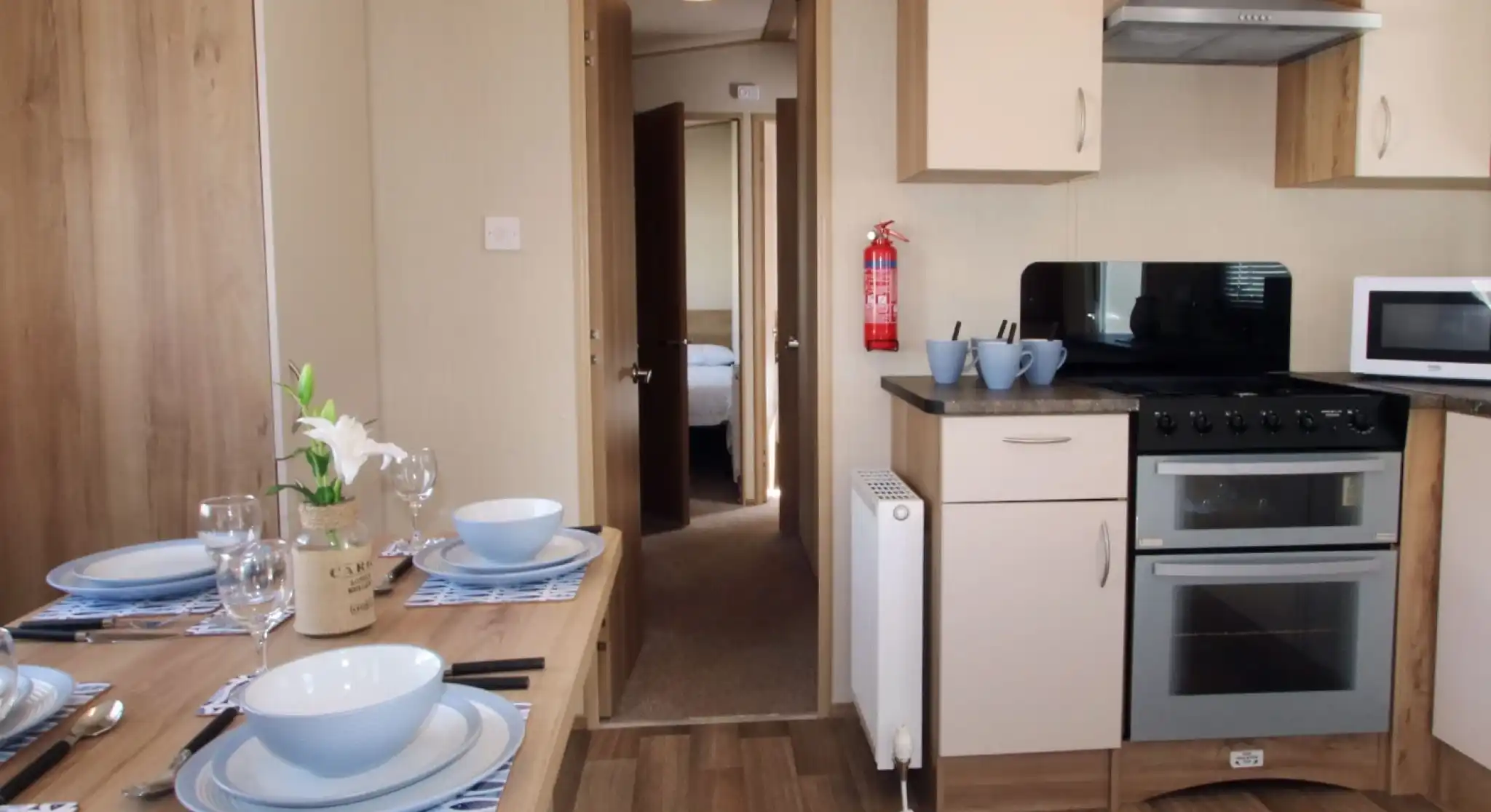 This image shows a clean, well-organized kitchen and dining area. The space is bright and functional, with light-colored cabinets and modern appliances, including an oven, microwave, and a fire extinguisher mounted on the wall. The dining table is set for a meal, with blue and white tableware and a simple vase with a flower adding a decorative touch. The flooring is wood-like laminate, contributing to a cozy and practical atmosphere. Through the open door, there’s a glimpse of a bedroom, showing a soft and comfortable environment.