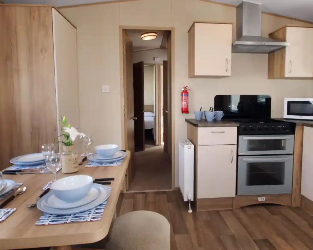 This image shows a clean, well-organized kitchen and dining area. The space is bright and functional, with light-colored cabinets and modern appliances, including an oven, microwave, and a fire extinguisher mounted on the wall. The dining table is set for a meal, with blue and white tableware and a simple vase with a flower adding a decorative touch. The flooring is wood-like laminate, contributing to a cozy and practical atmosphere. Through the open door, there’s a glimpse of a bedroom, showing a soft and comfortable environment.
