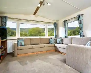 The image shows a cozy and well-lit living area, featuring a sectional sofa with soft, neutral tones and geometric accent pillows. The room has large windows offering a scenic view of a lush landscape with rolling hills, trees, and a clear sky. The space feels open and airy, with modern curtains that complement the sofa, creating a calm and inviting atmosphere. There’s also a TV unit in the corner, adding to the comfortable, relaxed vibe of the room. The soft carpet and wood elements enhance the homely, rustic charm.