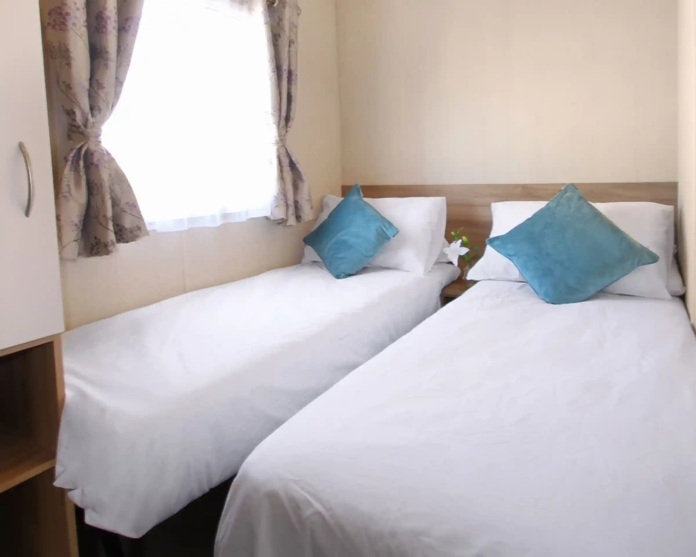 This image features a cozy twin bedroom with two single beds neatly arranged. Each bed is adorned with white linens and soft teal throw pillows, creating a fresh and inviting feel. The room has natural light coming through a window, with delicate floral curtains adding a touch of softness to the space. There’s a small plant on the bedside, adding a bit of greenery to the serene setting. The neutral tones and clean design give the room a calming and comfortable atmosphere.