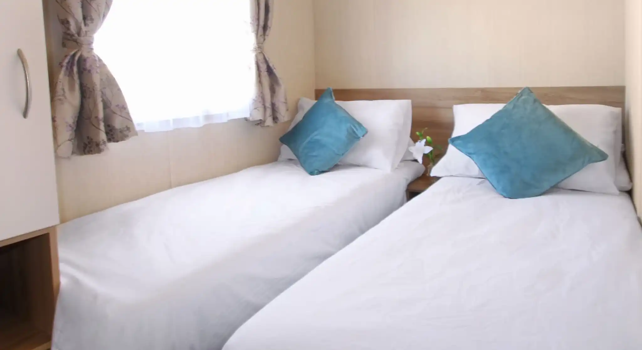 This image features a cozy twin bedroom with two single beds neatly arranged. Each bed is adorned with white linens and soft teal throw pillows, creating a fresh and inviting feel. The room has natural light coming through a window, with delicate floral curtains adding a touch of softness to the space. There’s a small plant on the bedside, adding a bit of greenery to the serene setting. The neutral tones and clean design give the room a calming and comfortable atmosphere.