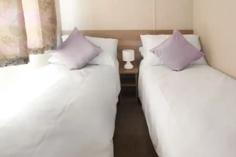 This image depicts another twin bedroom, similarly designed with a clean and simple aesthetic. The beds are neatly made with white linens and complemented by soft lavender-colored throw pillows. A small bedside table with a lamp sits between the beds, adding a cozy and functional touch. The room is bathed in natural light coming through a window with soft, patterned curtains. The neutral tones throughout the room contribute to a tranquil and inviting atmosphere.