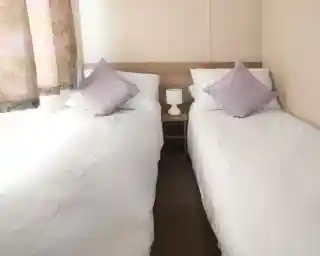 This image depicts another twin bedroom, similarly designed with a clean and simple aesthetic. The beds are neatly made with white linens and complemented by soft lavender-colored throw pillows. A small bedside table with a lamp sits between the beds, adding a cozy and functional touch. The room is bathed in natural light coming through a window with soft, patterned curtains. The neutral tones throughout the room contribute to a tranquil and inviting atmosphere.