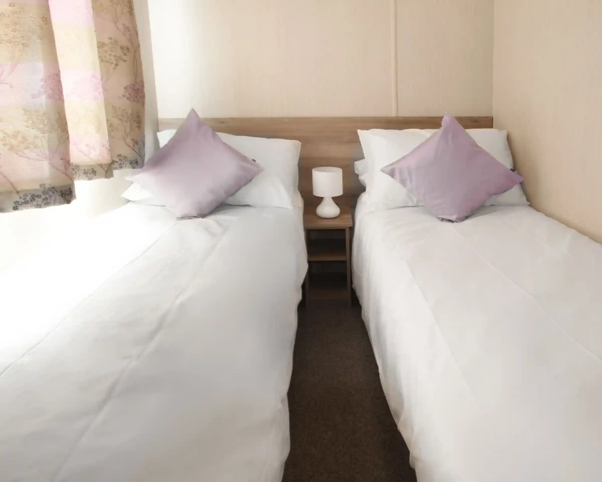 This image depicts another twin bedroom, similarly designed with a clean and simple aesthetic. The beds are neatly made with white linens and complemented by soft lavender-colored throw pillows. A small bedside table with a lamp sits between the beds, adding a cozy and functional touch. The room is bathed in natural light coming through a window with soft, patterned curtains. The neutral tones throughout the room contribute to a tranquil and inviting atmosphere.