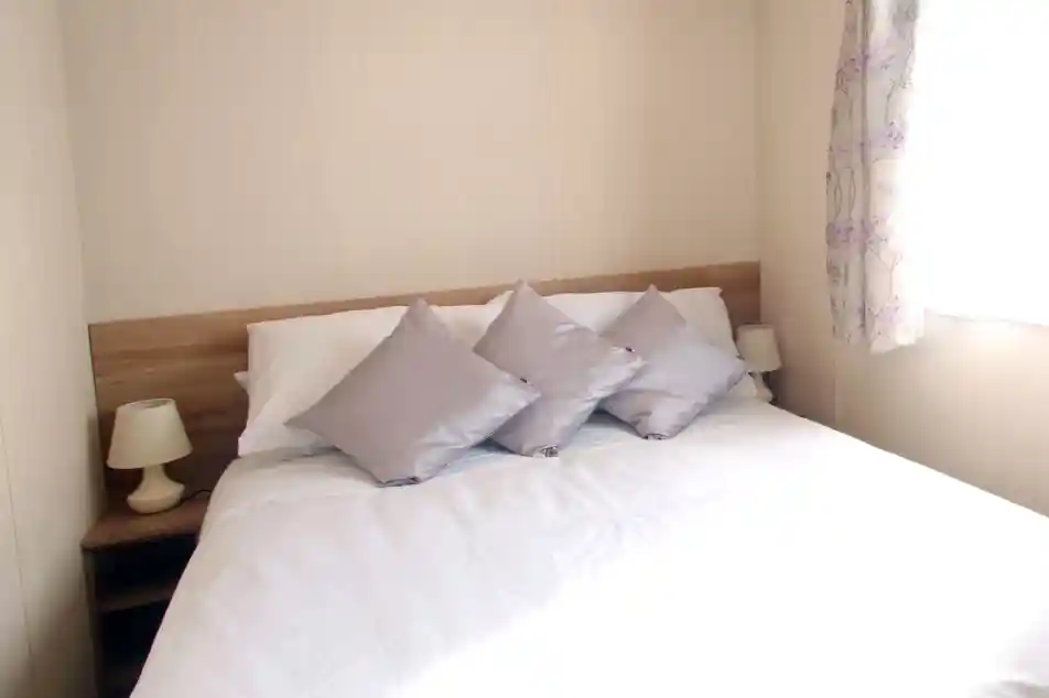 This image shows a serene and cozy master bedroom with a simple yet elegant design. The bed features white linens with soft silver-gray pillows, creating a calm and inviting atmosphere. Two small lamps sit on either side of the bed, complementing the light wood bedside tables. The room is bathed in natural light from the window, which is adorned with soft floral curtains, adding a touch of color. The overall ambiance is peaceful, with neutral tones and a clean, uncluttered aesthetic.