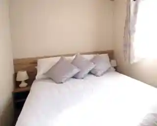 This image shows a serene and cozy master bedroom with a simple yet elegant design. The bed features white linens with soft silver-gray pillows, creating a calm and inviting atmosphere. Two small lamps sit on either side of the bed, complementing the light wood bedside tables. The room is bathed in natural light from the window, which is adorned with soft floral curtains, adding a touch of color. The overall ambiance is peaceful, with neutral tones and a clean, uncluttered aesthetic.