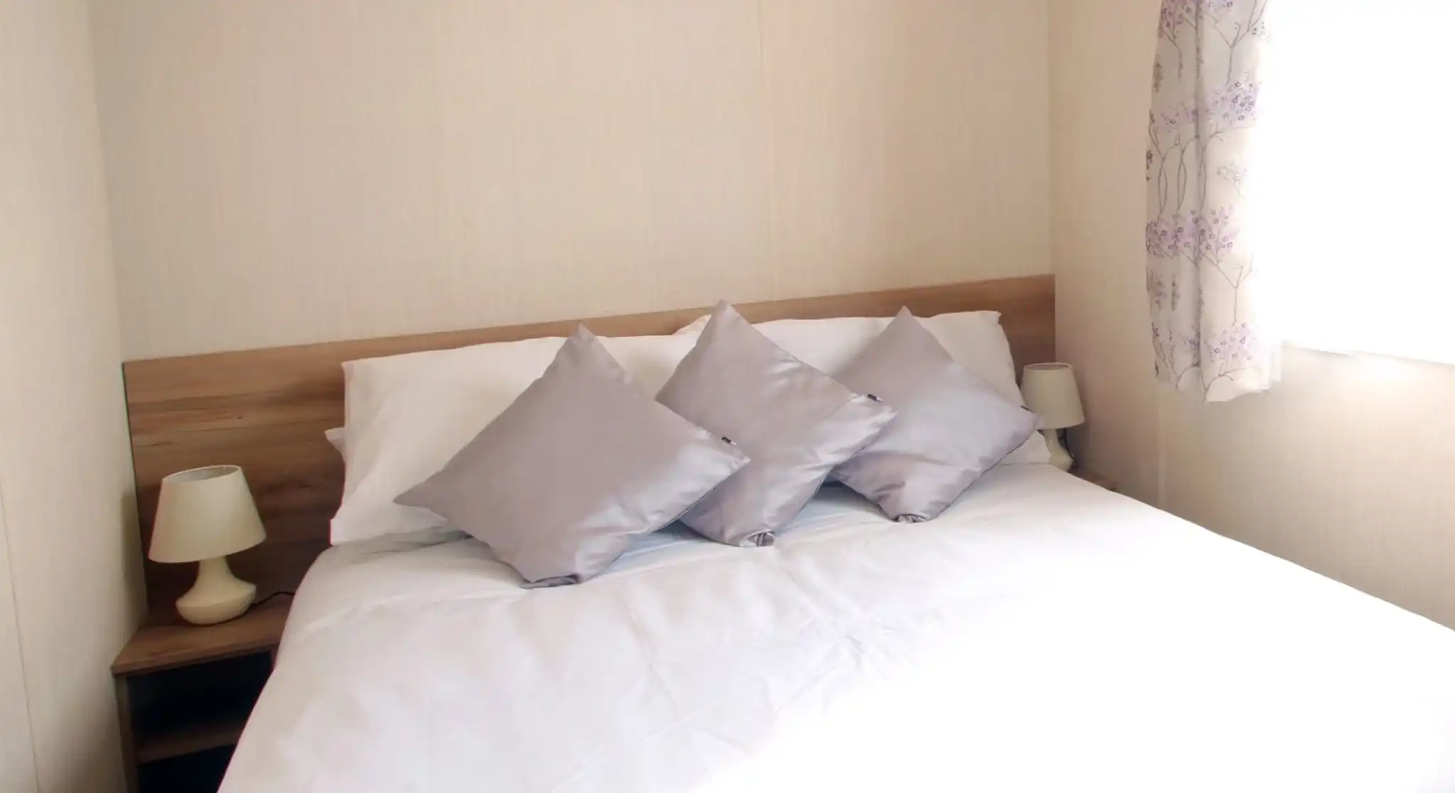 This image shows a serene and cozy master bedroom with a simple yet elegant design. The bed features white linens with soft silver-gray pillows, creating a calm and inviting atmosphere. Two small lamps sit on either side of the bed, complementing the light wood bedside tables. The room is bathed in natural light from the window, which is adorned with soft floral curtains, adding a touch of color. The overall ambiance is peaceful, with neutral tones and a clean, uncluttered aesthetic.