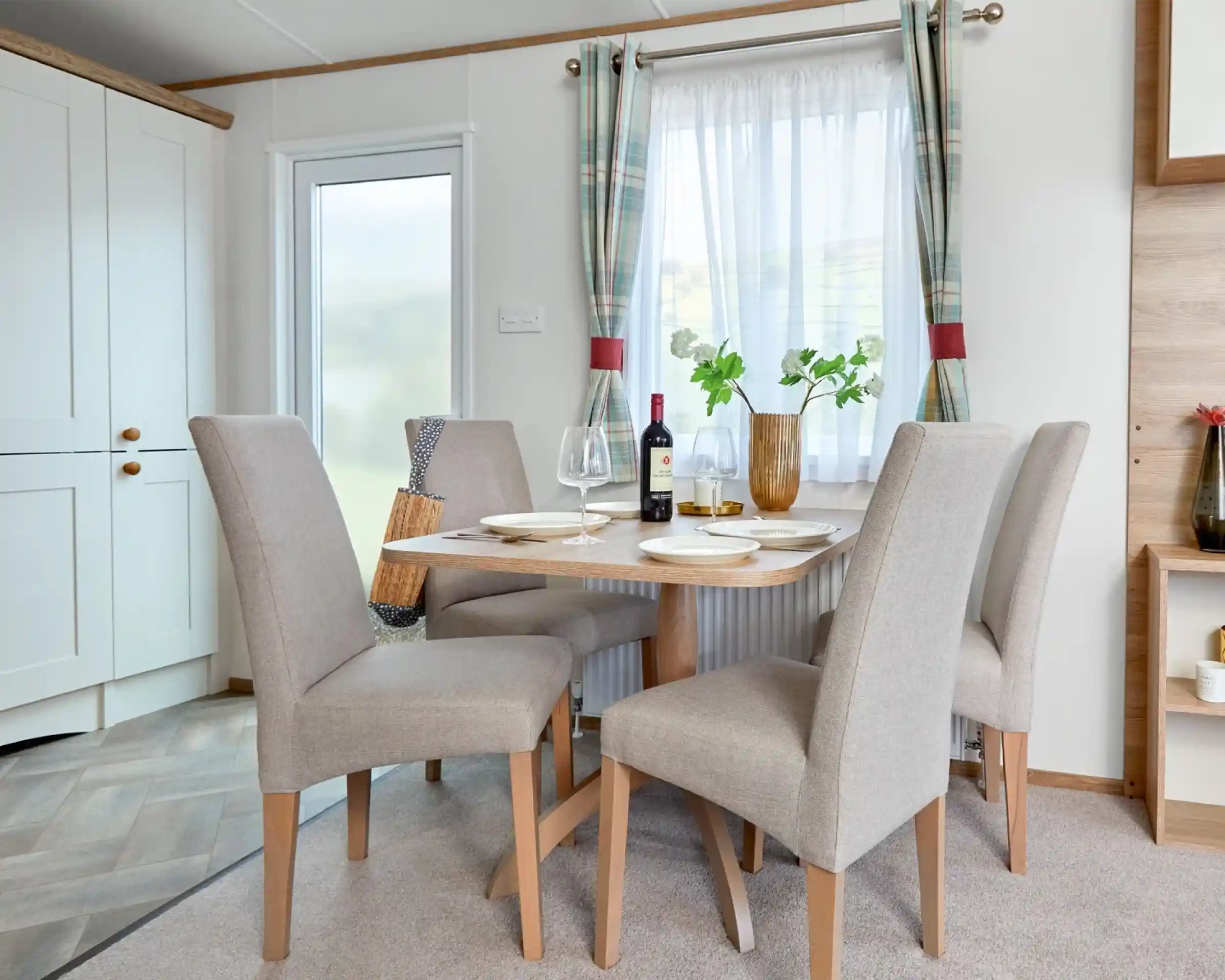 Modern dining area inside the ABI Wimbledon holiday home, featuring a wooden table set for four with beige upholstered chairs, a bottle of wine, and a decorative vase with greenery. The space is bright with natural light coming through a window dressed with plaid curtains, and a frosted glass door and light cabinetry are visible in the background.