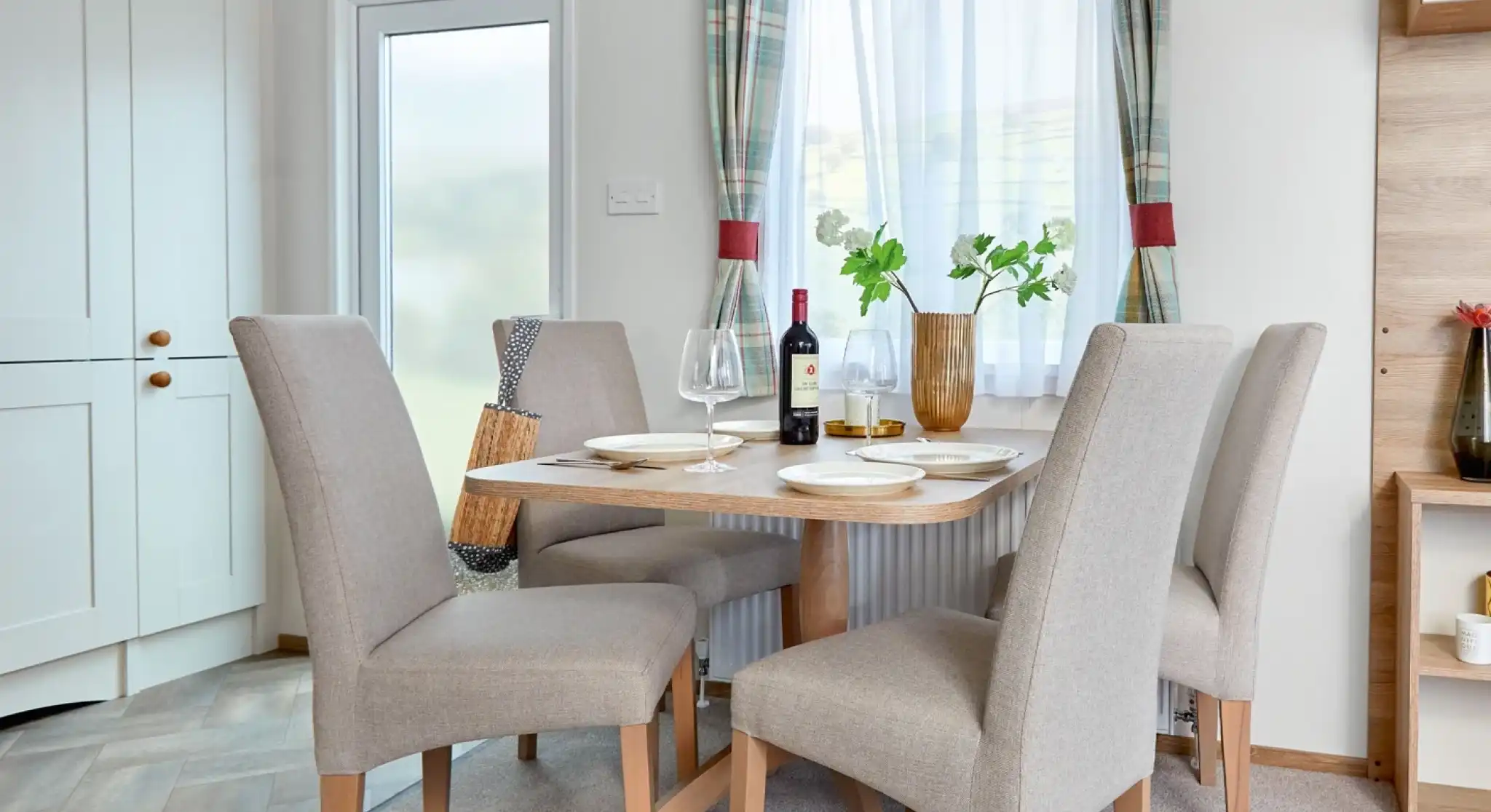 Modern dining area inside the ABI Wimbledon holiday home, featuring a wooden table set for four with beige upholstered chairs, a bottle of wine, and a decorative vase with greenery. The space is bright with natural light coming through a window dressed with plaid curtains, and a frosted glass door and light cabinetry are visible in the background.