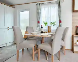 Modern dining area inside the ABI Wimbledon holiday home, featuring a wooden table set for four with beige upholstered chairs, a bottle of wine, and a decorative vase with greenery. The space is bright with natural light coming through a window dressed with plaid curtains, and a frosted glass door and light cabinetry are visible in the background.