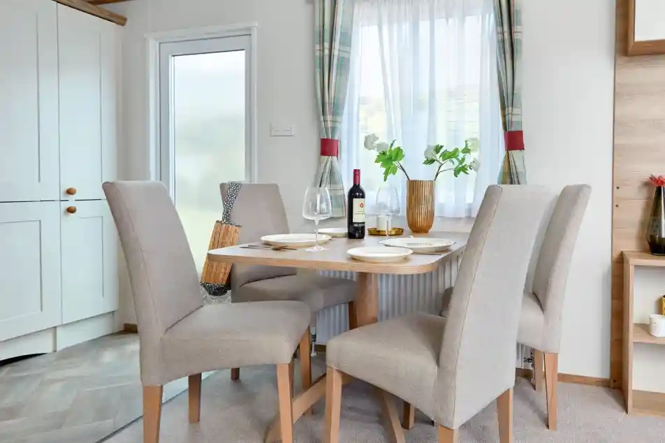 Modern dining area inside the ABI Wimbledon holiday home, featuring a wooden table set for four with beige upholstered chairs, a bottle of wine, and a decorative vase with greenery. The space is bright with natural light coming through a window dressed with plaid curtains, and a frosted glass door and light cabinetry are visible in the background.