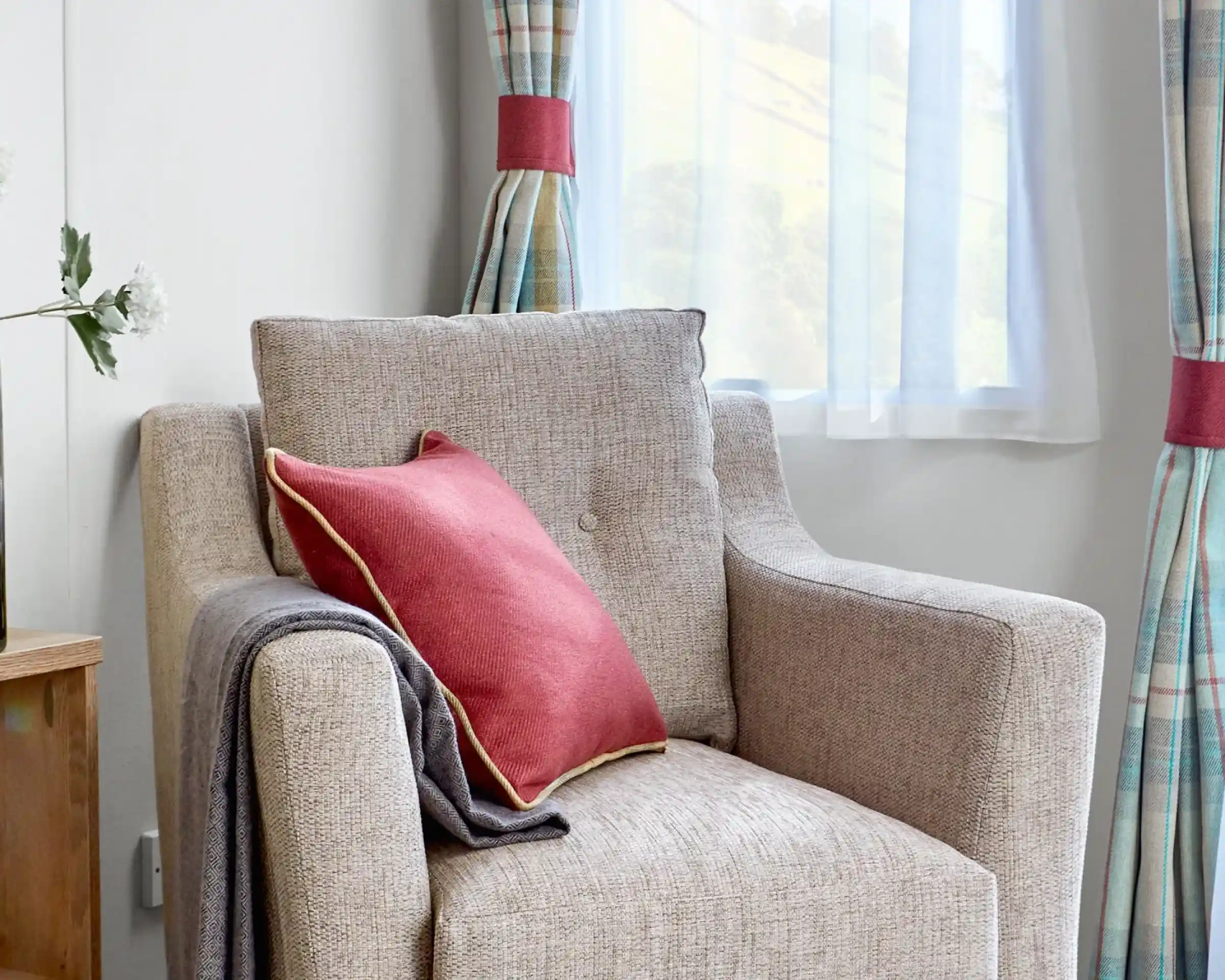Comfortable beige fabric armchair inside the ABI Wimbledon holiday home, styled with a red cushion featuring gold piping and a neatly draped grey throw. The chair is positioned near a window with sheer white curtains and tartan drapes, allowing soft natural light to fill the space.