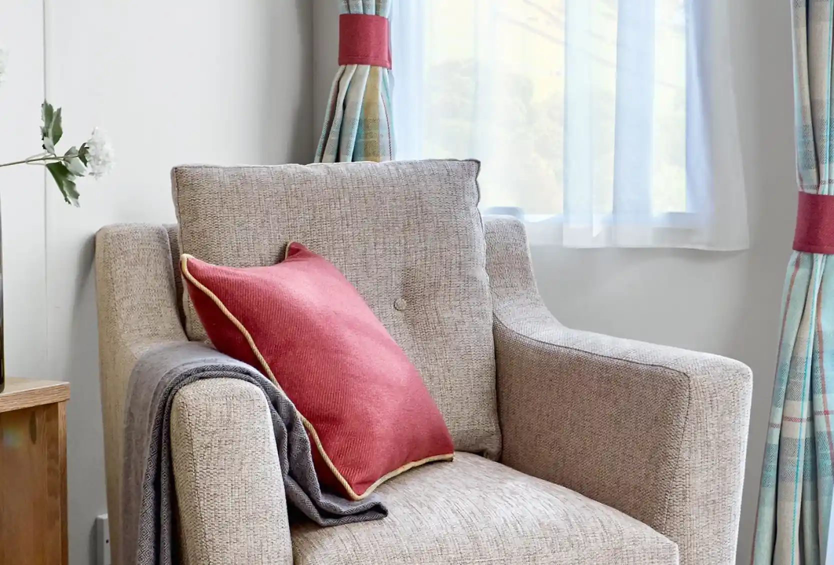 Comfortable beige fabric armchair inside the ABI Wimbledon holiday home, styled with a red cushion featuring gold piping and a neatly draped grey throw. The chair is positioned near a window with sheer white curtains and tartan drapes, allowing soft natural light to fill the space.