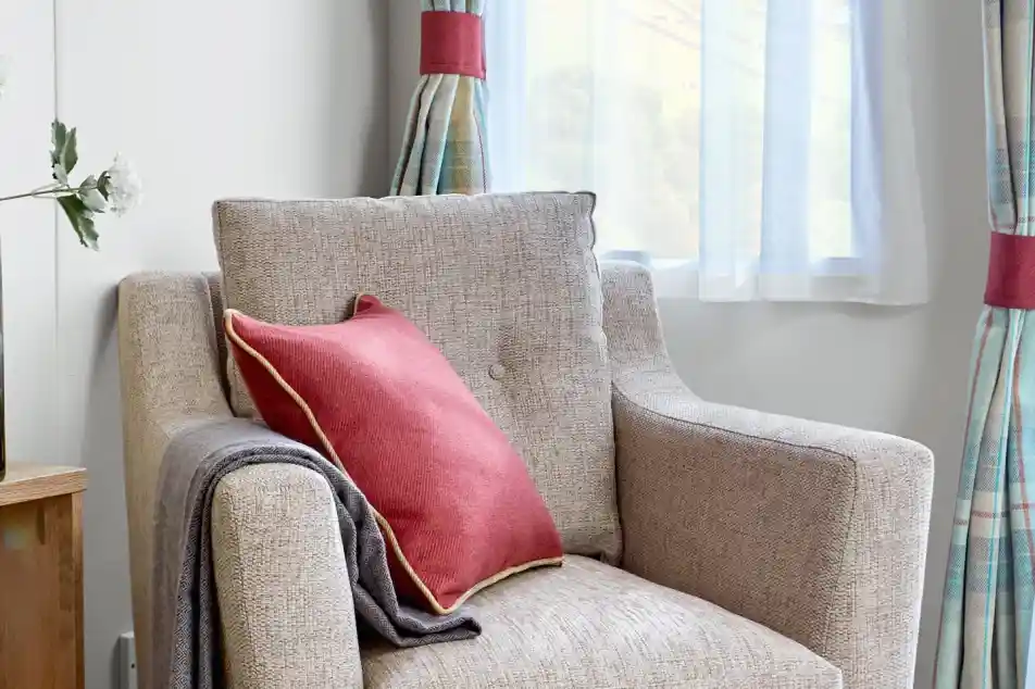 Comfortable beige fabric armchair inside the ABI Wimbledon holiday home, styled with a red cushion featuring gold piping and a neatly draped grey throw. The chair is positioned near a window with sheer white curtains and tartan drapes, allowing soft natural light to fill the space.