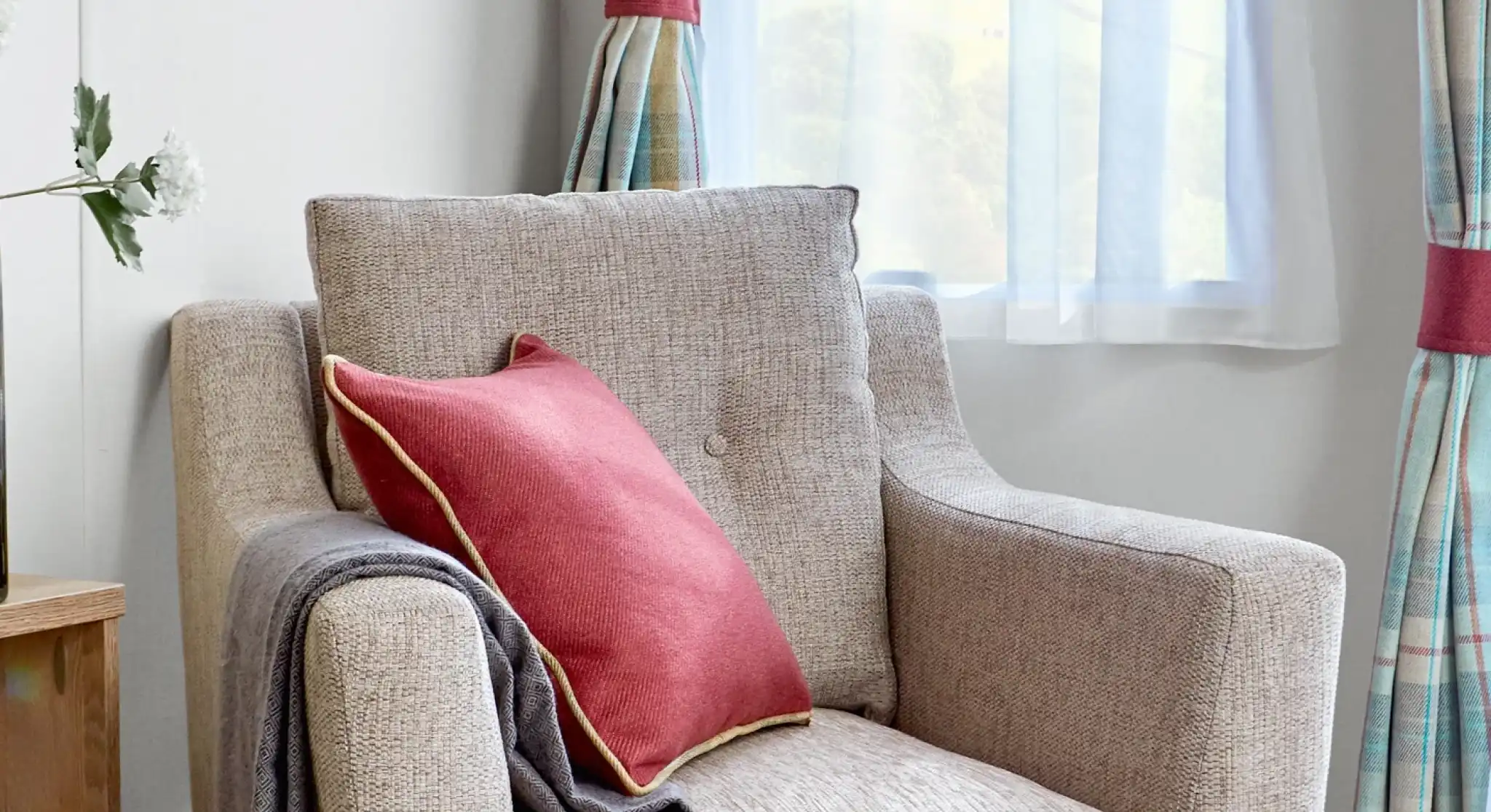 Comfortable beige fabric armchair inside the ABI Wimbledon holiday home, styled with a red cushion featuring gold piping and a neatly draped grey throw. The chair is positioned near a window with sheer white curtains and tartan drapes, allowing soft natural light to fill the space.