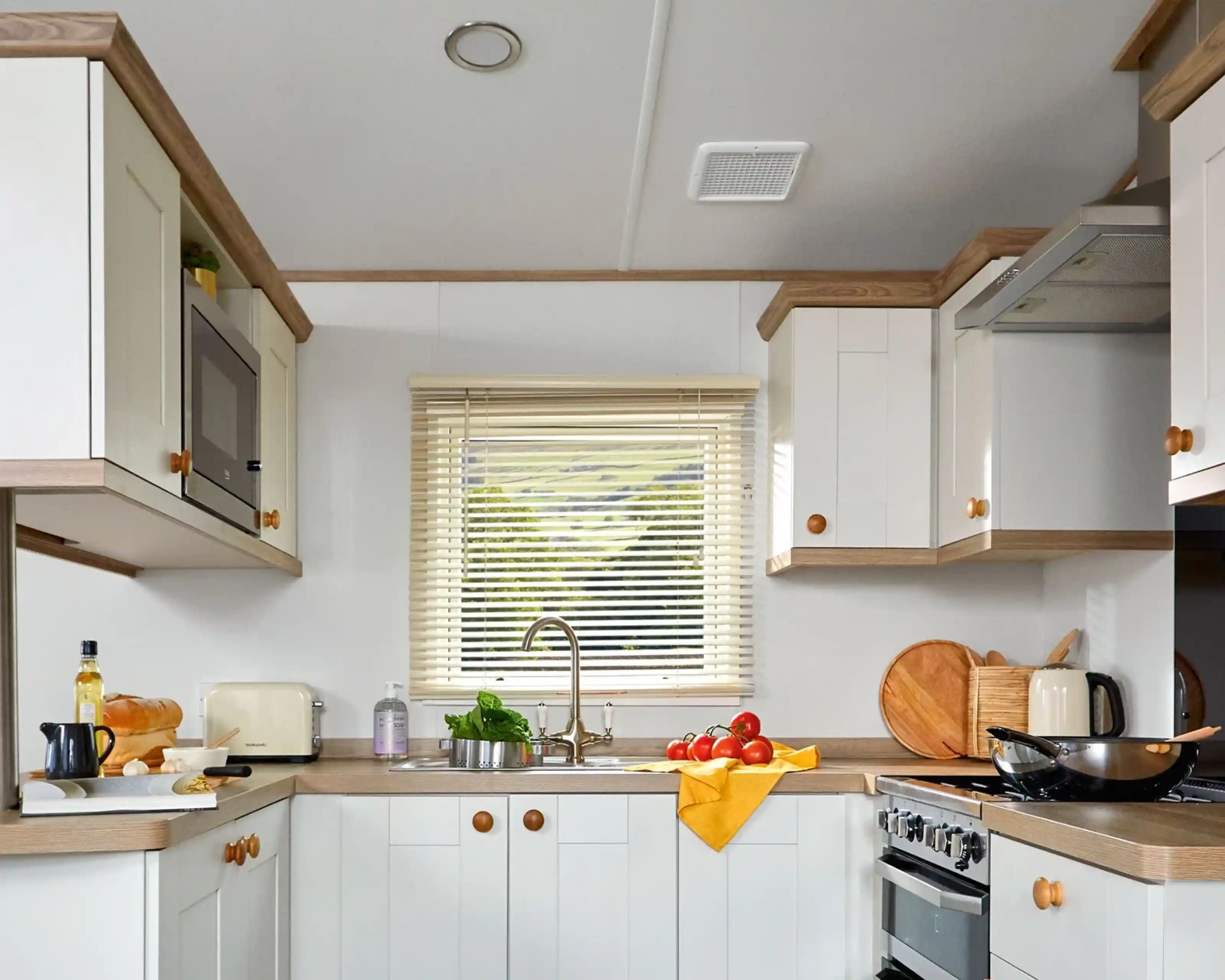 This image showcases a cozy and functional kitchen with a light and bright color scheme. The cabinets are a mix of white with wooden accents, creating a warm and inviting atmosphere. The kitchen features essential appliances, including a microwave, toaster, and kettle. The counter is neatly arranged with some ingredients for cooking, such as tomatoes, a loaf of bread, olive oil, and a cutting board. The sink is positioned below a window, allowing natural light to illuminate the space and offering a view of the outdoors. The overall design is clean, practical, and comfortable for cooking and dining.