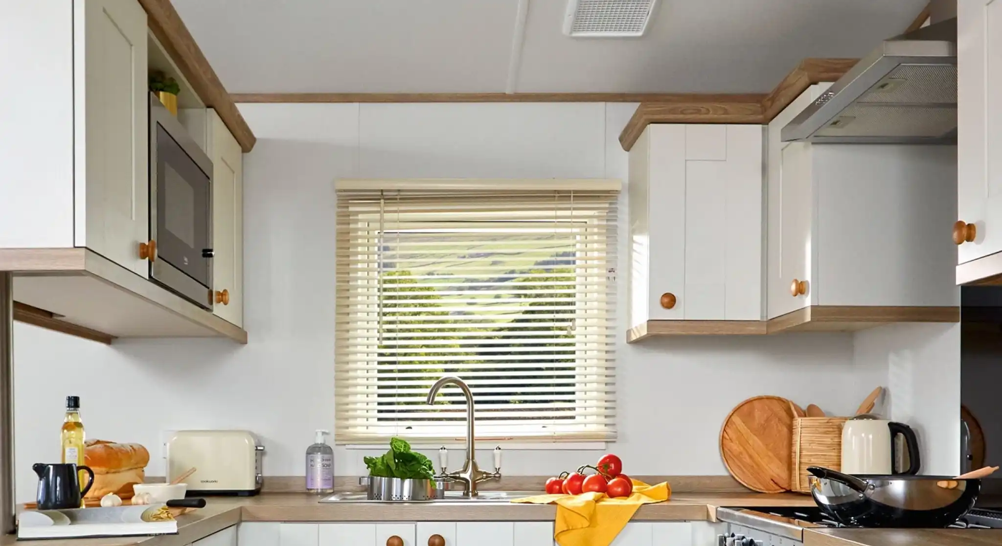 This image showcases a cozy and functional kitchen with a light and bright color scheme. The cabinets are a mix of white with wooden accents, creating a warm and inviting atmosphere. The kitchen features essential appliances, including a microwave, toaster, and kettle. The counter is neatly arranged with some ingredients for cooking, such as tomatoes, a loaf of bread, olive oil, and a cutting board. The sink is positioned below a window, allowing natural light to illuminate the space and offering a view of the outdoors. The overall design is clean, practical, and comfortable for cooking and dining.
