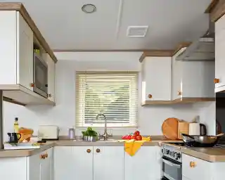 This image showcases a cozy and functional kitchen with a light and bright color scheme. The cabinets are a mix of white with wooden accents, creating a warm and inviting atmosphere. The kitchen features essential appliances, including a microwave, toaster, and kettle. The counter is neatly arranged with some ingredients for cooking, such as tomatoes, a loaf of bread, olive oil, and a cutting board. The sink is positioned below a window, allowing natural light to illuminate the space and offering a view of the outdoors. The overall design is clean, practical, and comfortable for cooking and dining.