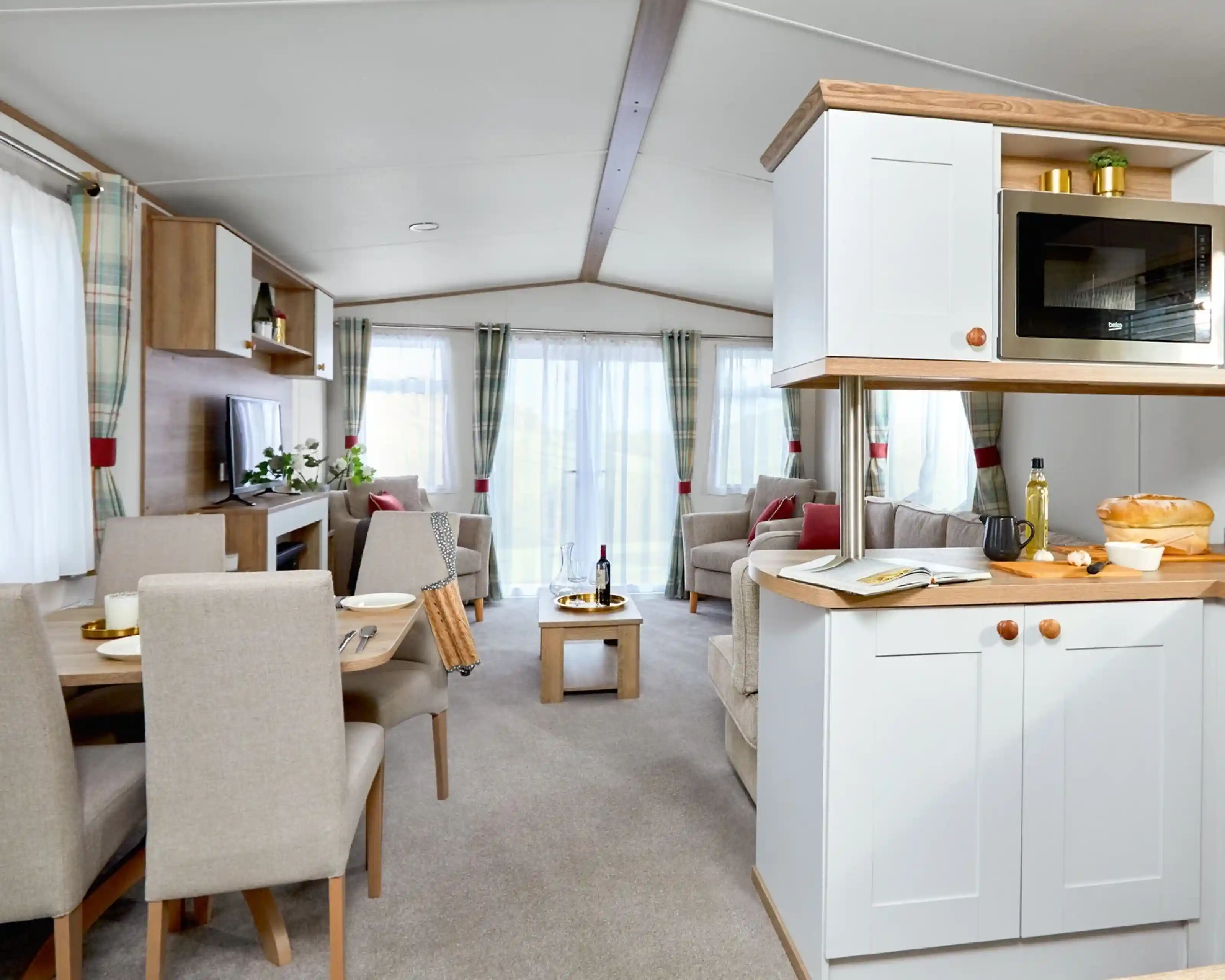 Bright and airy open-plan lounge, kitchen, and dining area inside the ABI Wimbledon holiday home. The space features neutral-toned furnishings, including a dining table with four upholstered chairs, an L-shaped sofa, an armchair, and a wooden coffee table. Large windows with sheer white curtains and plaid drapes let in plenty of natural light. The kitchen includes white cabinetry, a built-in microwave, and wooden countertops with fresh bread and olive oil on display. A TV and shelving unit complete the cozy yet modern interior.