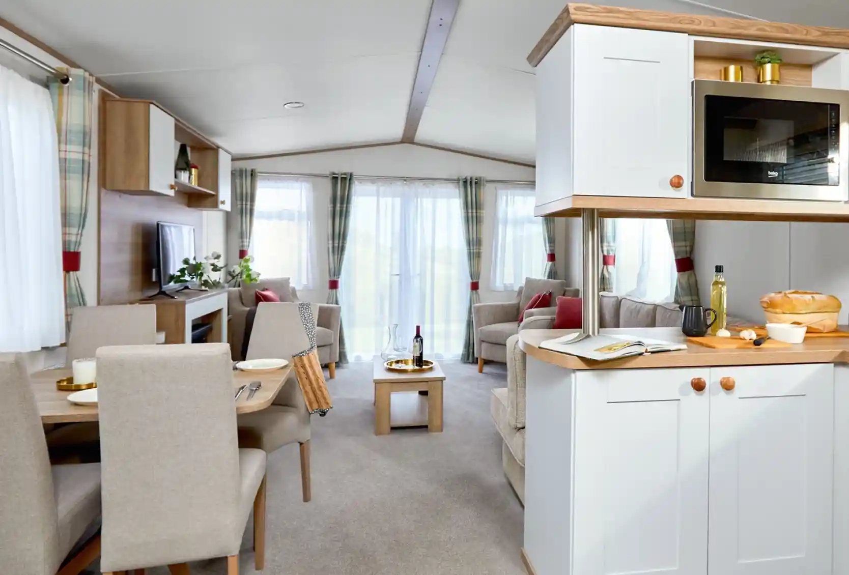 Bright and airy open-plan lounge, kitchen, and dining area inside the ABI Wimbledon holiday home. The space features neutral-toned furnishings, including a dining table with four upholstered chairs, an L-shaped sofa, an armchair, and a wooden coffee table. Large windows with sheer white curtains and plaid drapes let in plenty of natural light. The kitchen includes white cabinetry, a built-in microwave, and wooden countertops with fresh bread and olive oil on display. A TV and shelving unit complete the cozy yet modern interior.